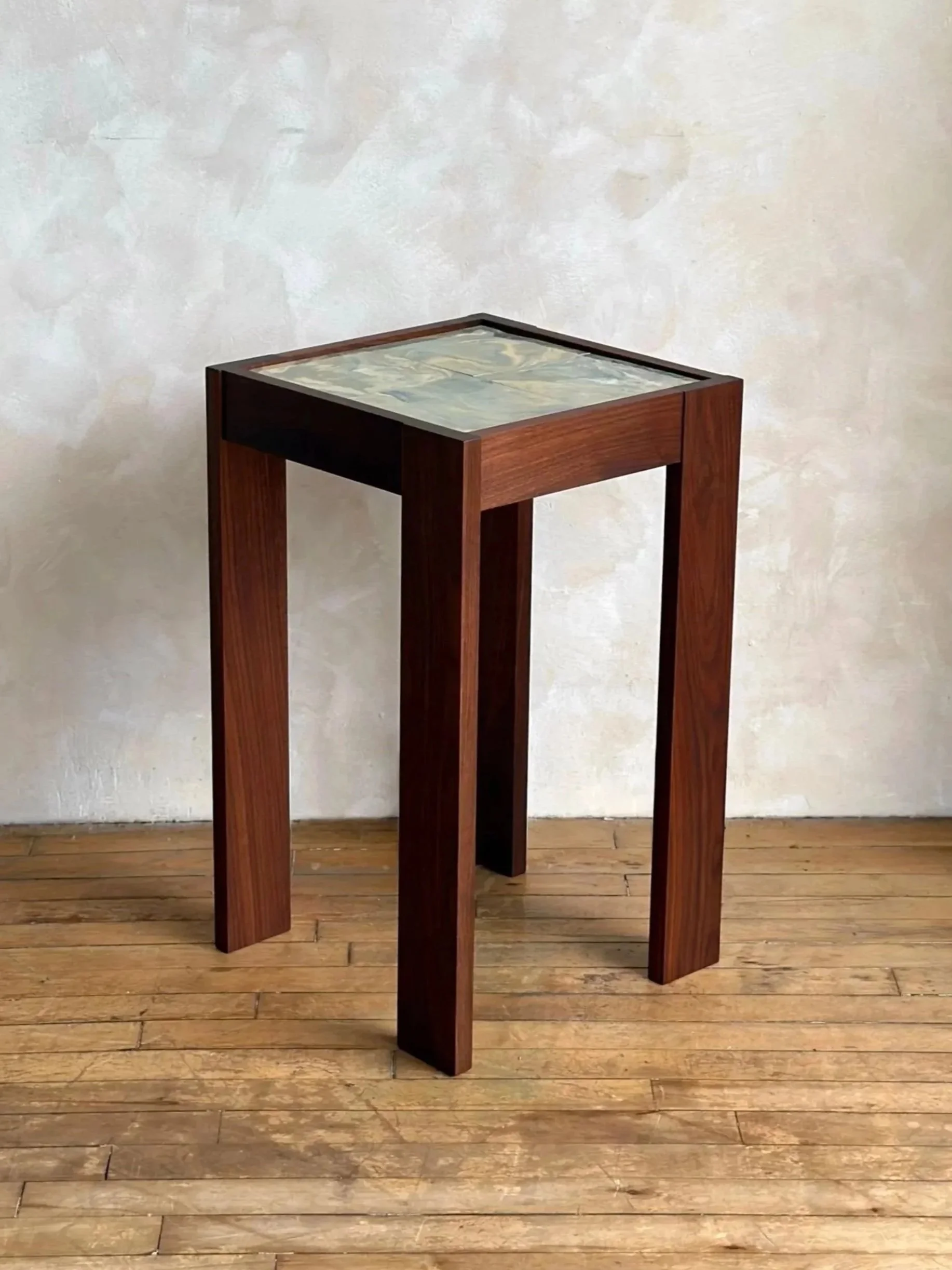 A wooden side table with a ceramic top, standing on a wooden floor against a plain wall.