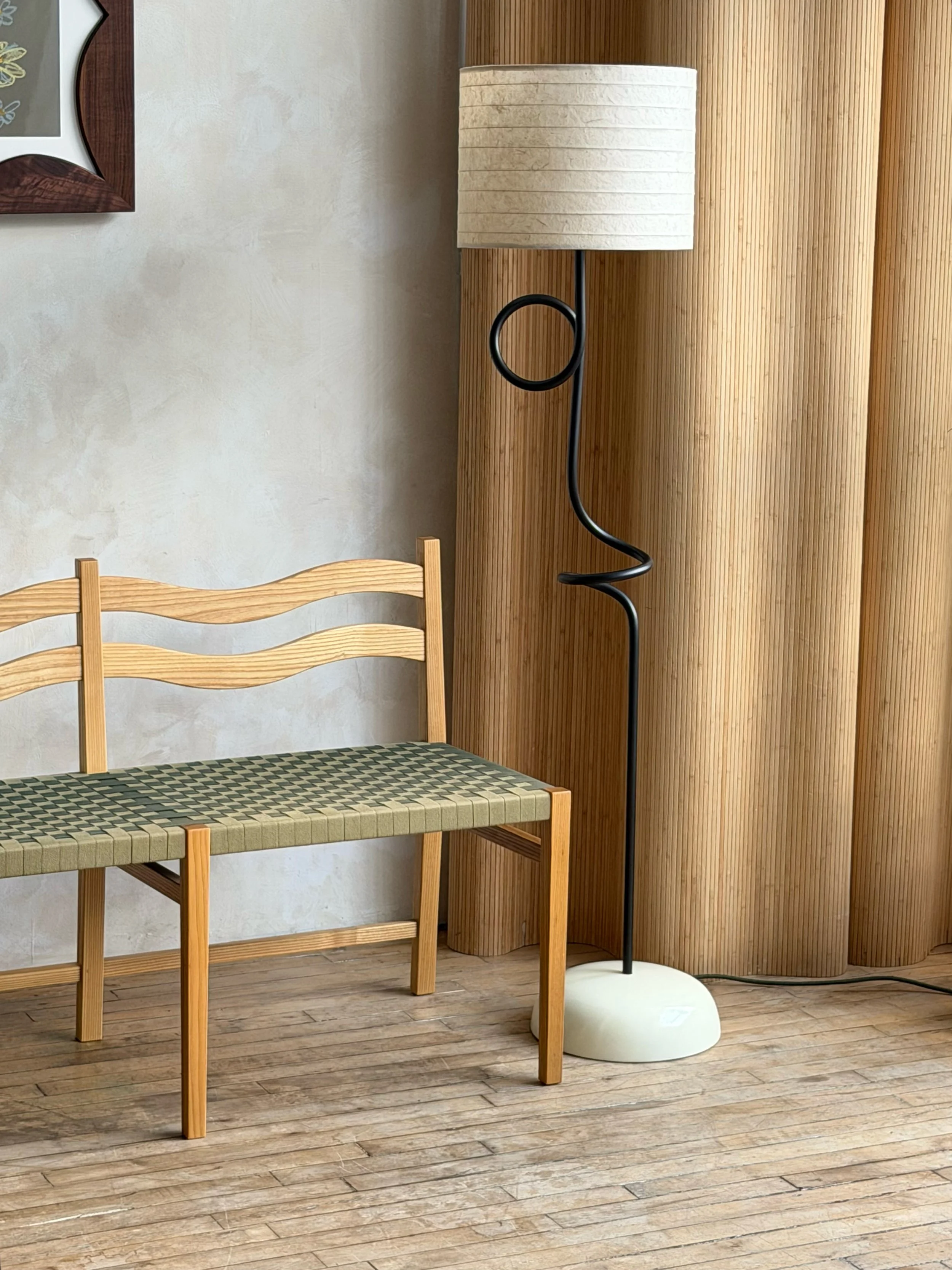 A wooden bench with a woven seat in front of a wall, next to a curved wooden room divider and a sculptural metal floor lamp with a paper shade.