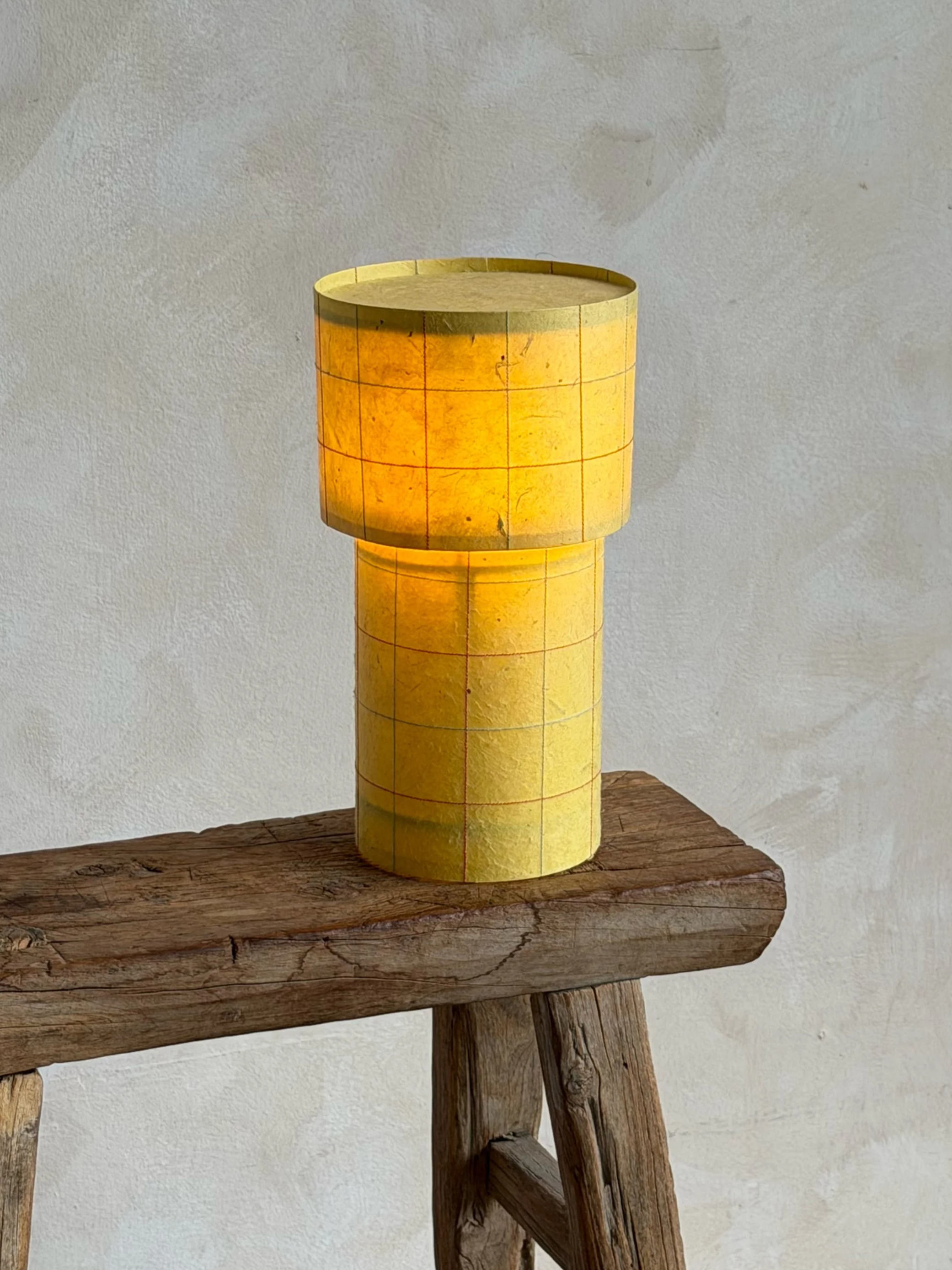 A yellow cylindrical lamp with a grid pattern, placed on a rustic wooden table against a white textured wall.