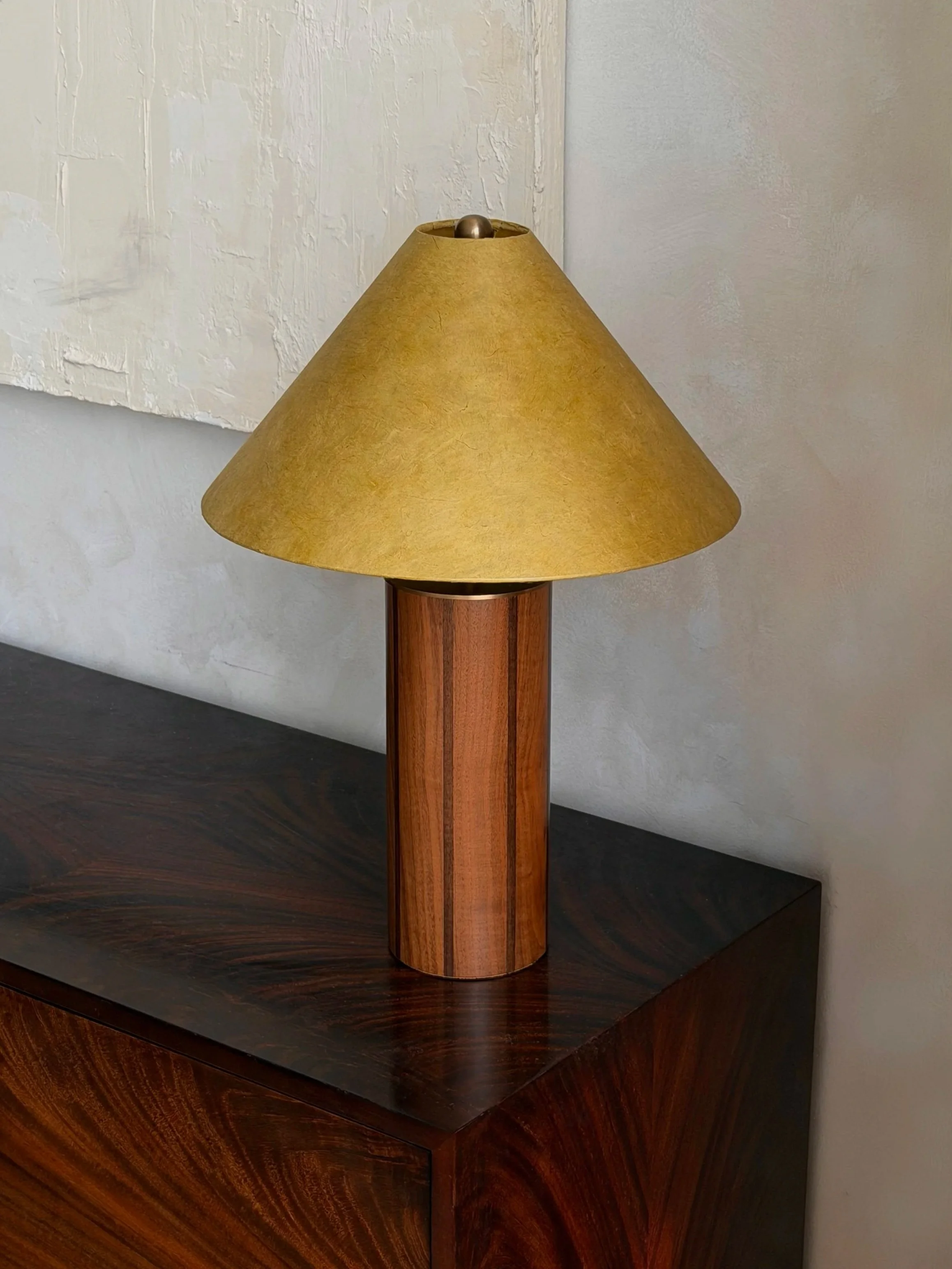A table lamp with a dark wooden base and a green lampshade placed on a wooden table against a textured wall.