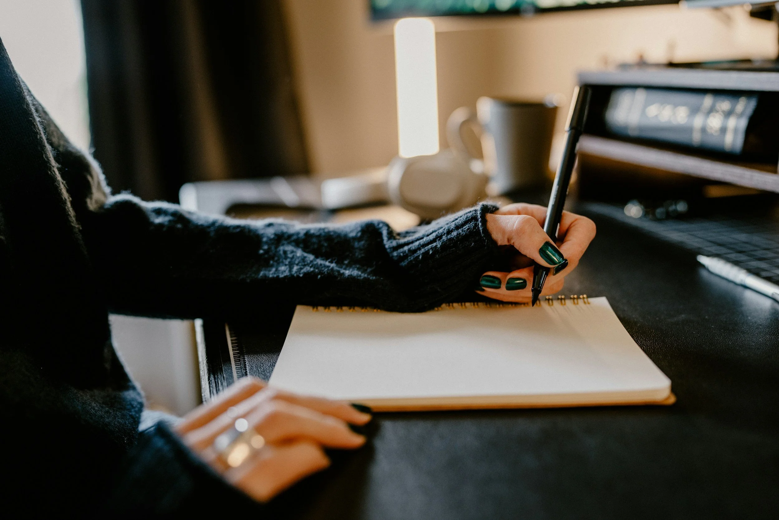 A person with dark green painted nails writing in a spiral notebook with a black pen at a desk. They are wearing a sleeve of a dark ribbed sweater and a wristwatch. In the background, there is a cup and a stack of papers or books on a desk, with sunlight coming through a window.