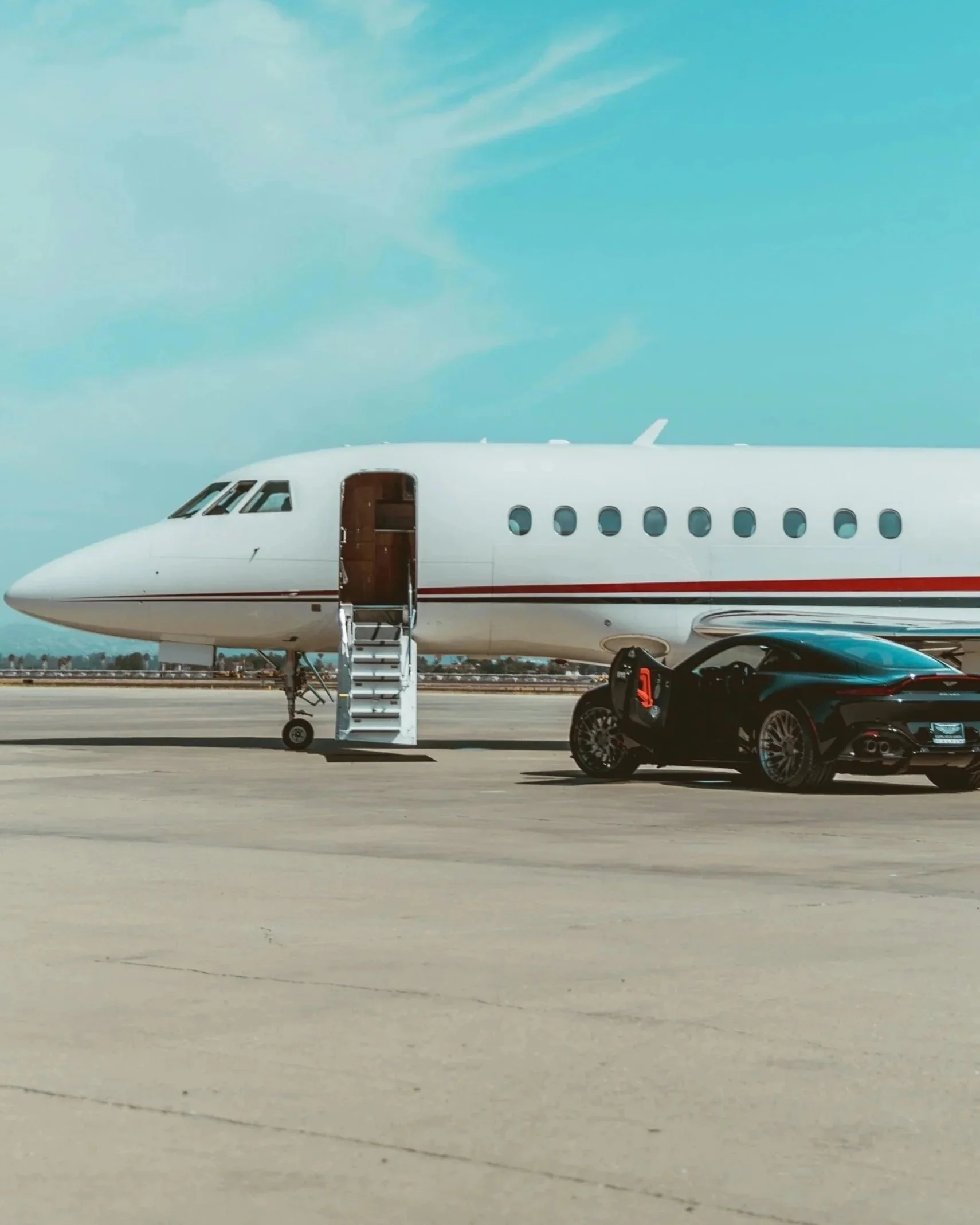 A white private jet airplane parked on the tarmac with an open door and stairs leading up to it. There is a black car parked nearby. The sky is partly cloudy.