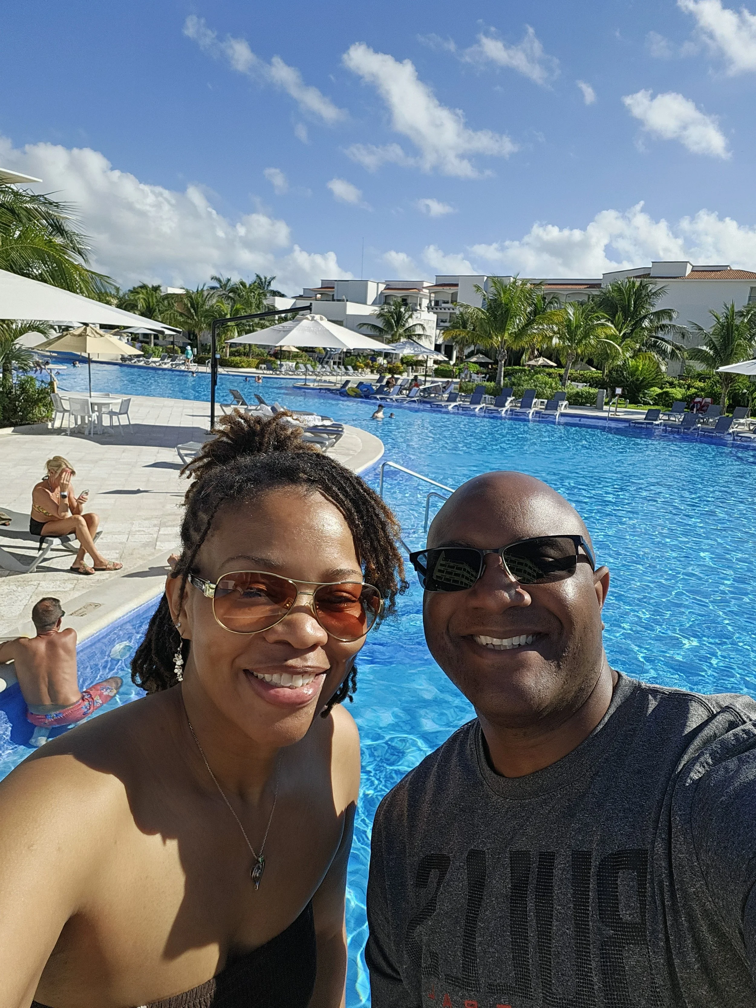 A smiling woman and man taking a selfie at a poolside resort with lounge chairs, umbrellas, palm trees, and buildings in the background on a sunny day.