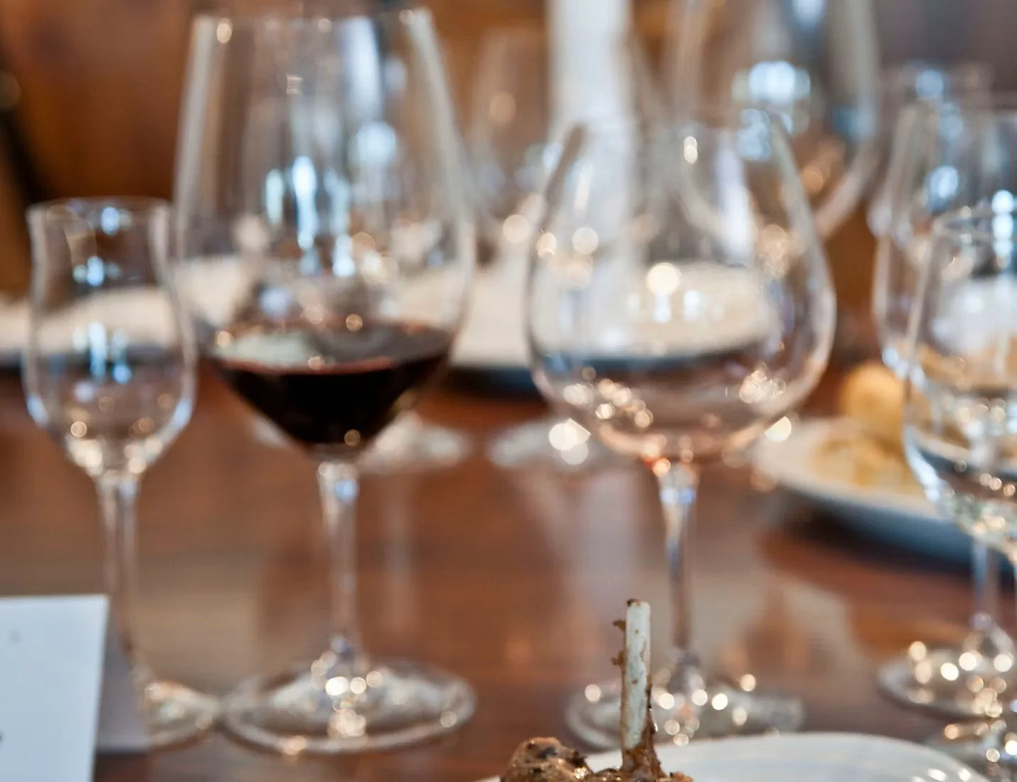 Multiple wine glasses on a wooden table, with one glass of red wine and some plates and utensils in the background.