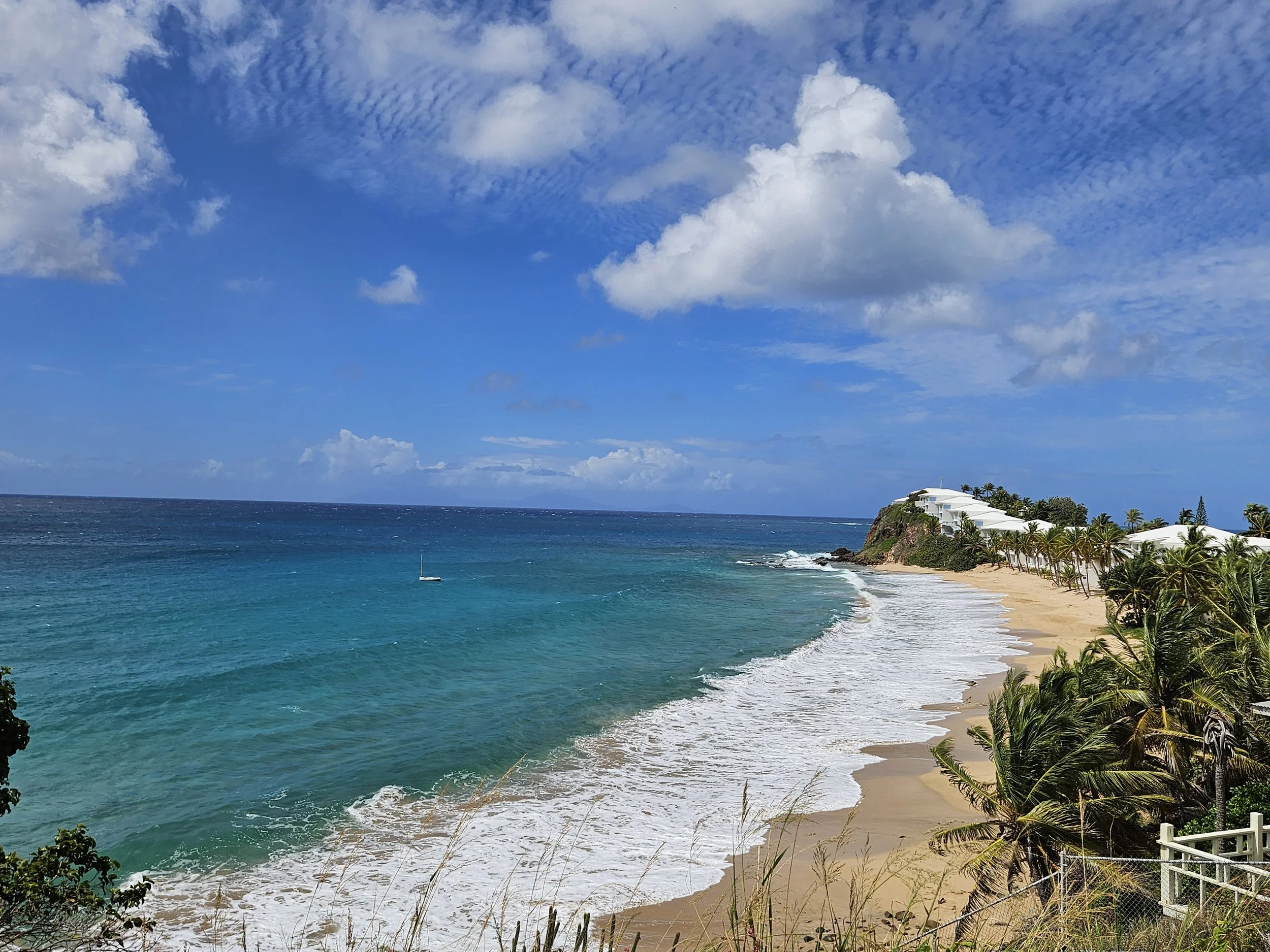 A beach scene with turquoise water, white sandy shore, lush palm trees, and a white building on a cliff overlooking the ocean.