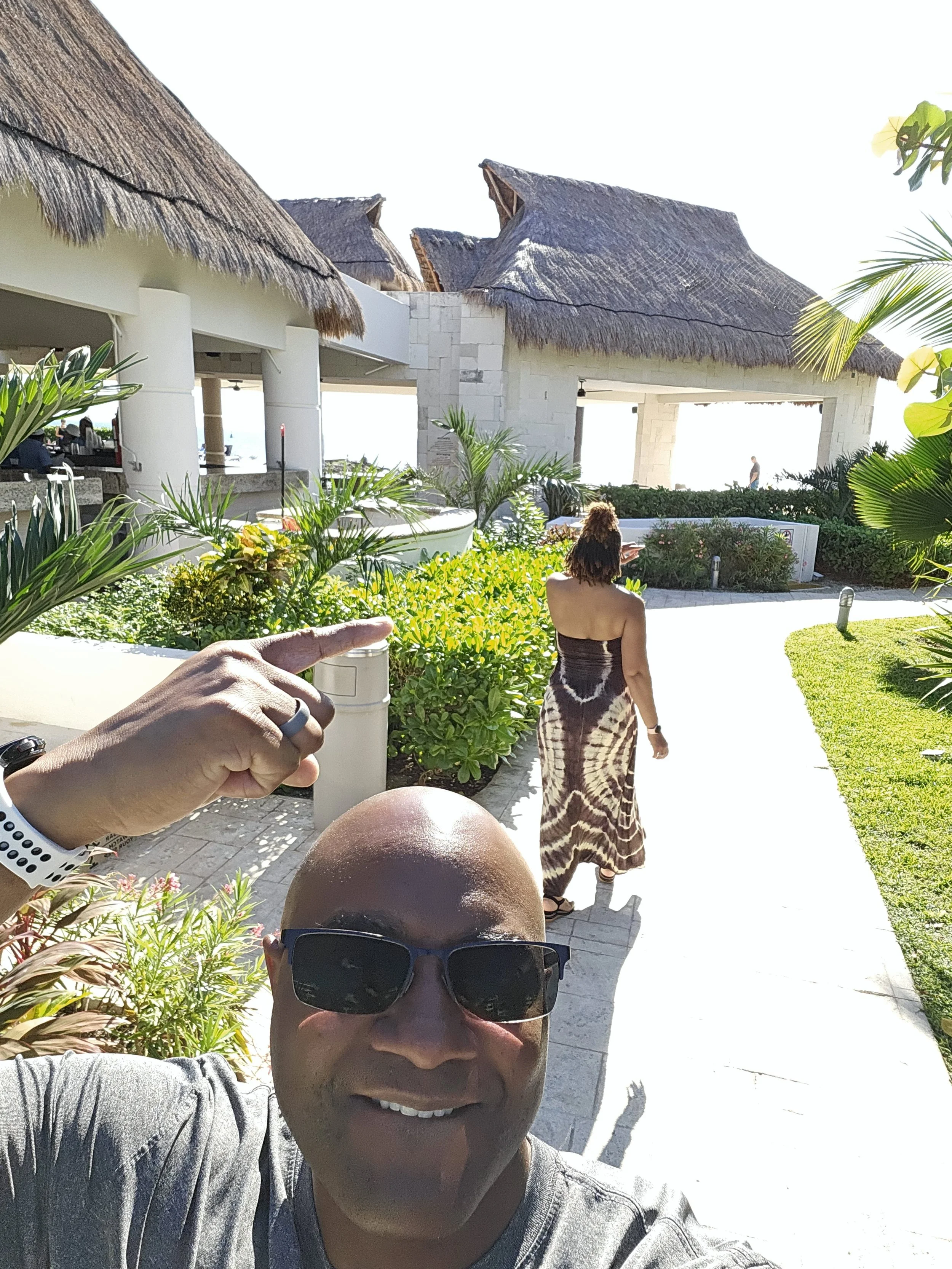 A man with dark glasses taking a selfie outdoors, with a woman walking away in the background, lush green plants, a white building with thatched roofs, and bright sunlight.
