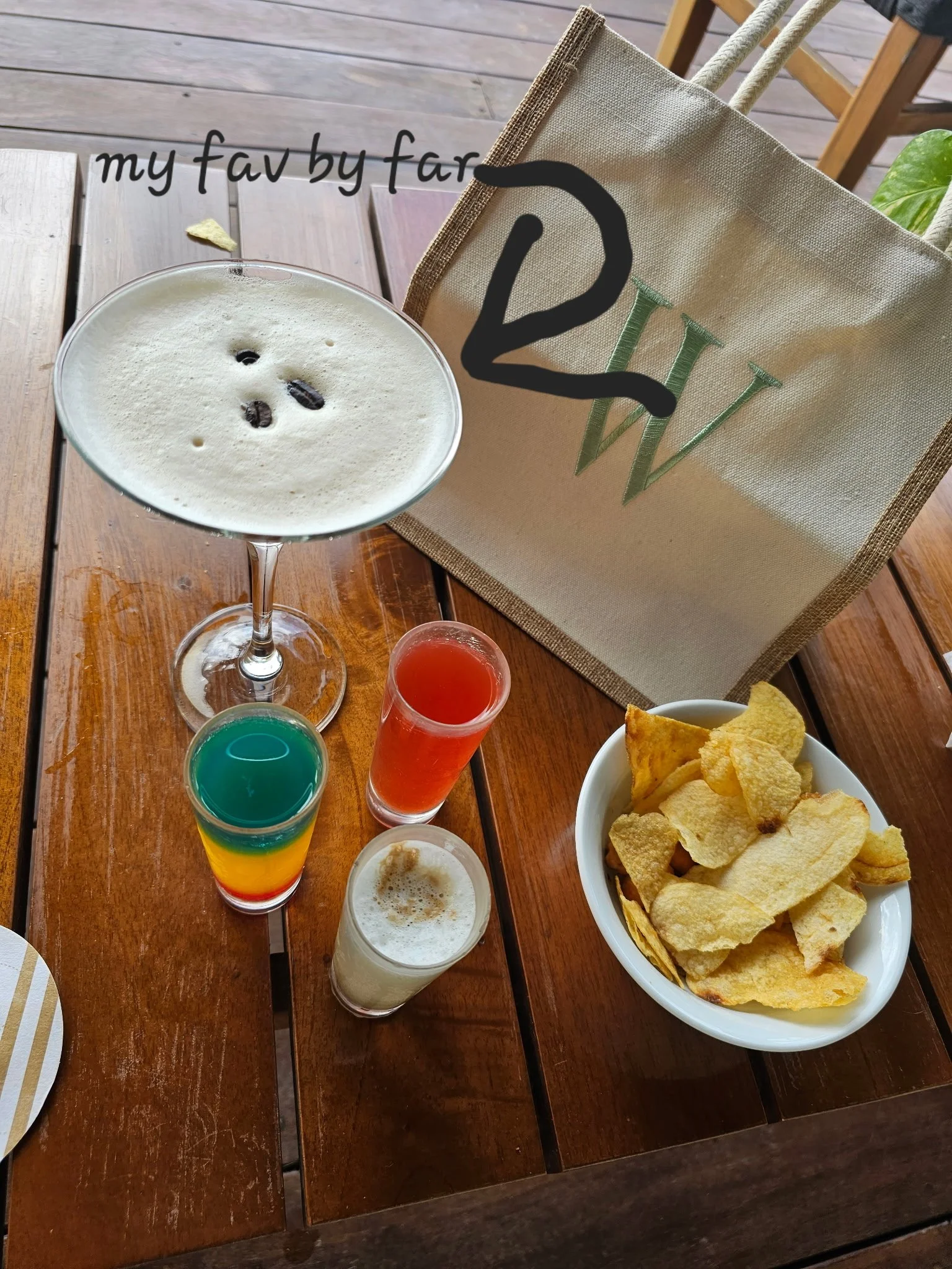 On a wooden table, there are several colorful drinks, a bowl of potato chips, and a glass of beer with foam. There is a beige bag with green and black embroidery, and the background shows part of a chair and outdoor flooring.