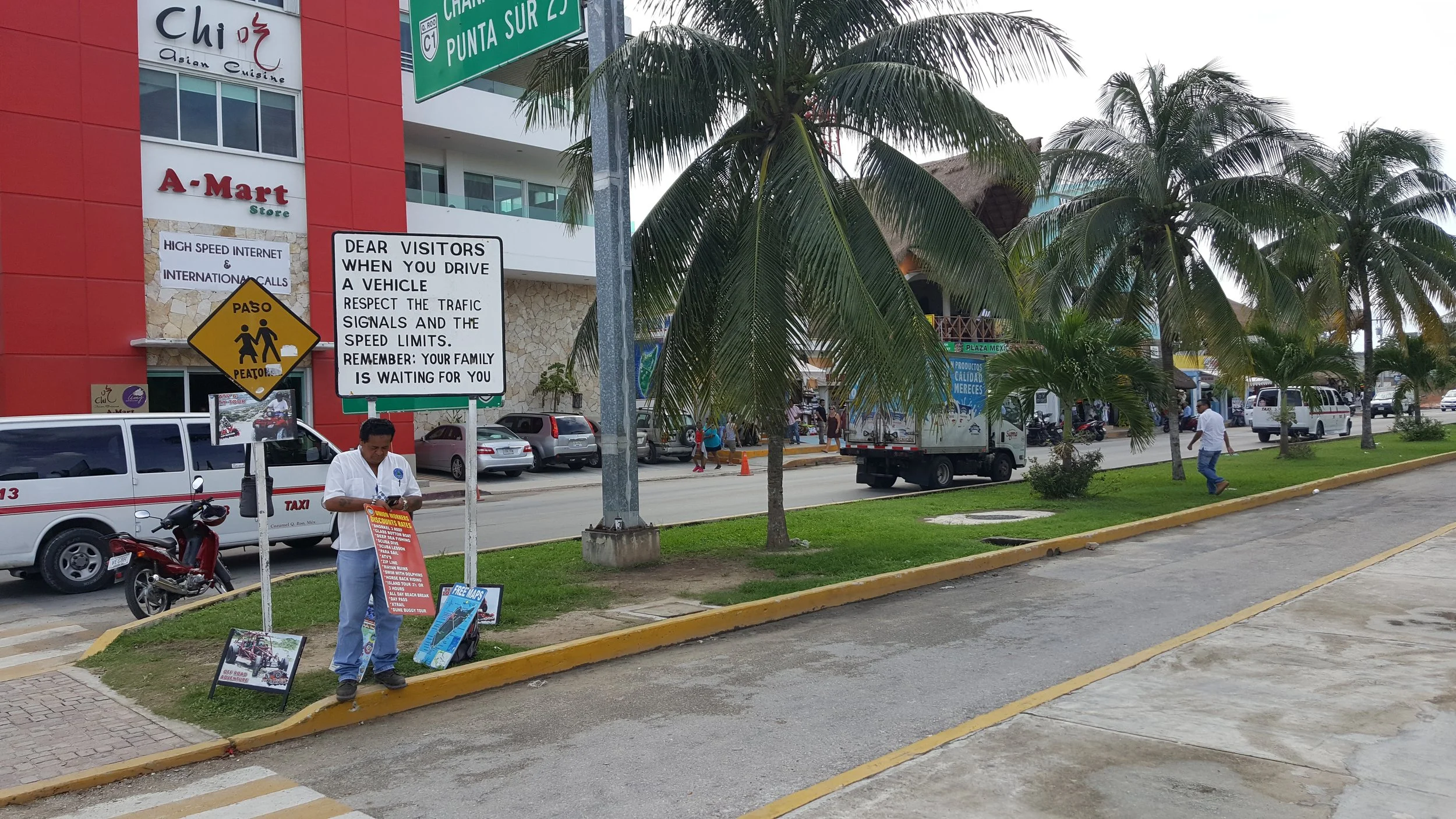 Street scene with palm trees, parked cars, and pedestrians, with signs including traffic instructions and a building with signs for an Asian cuisine restaurant and a store.
