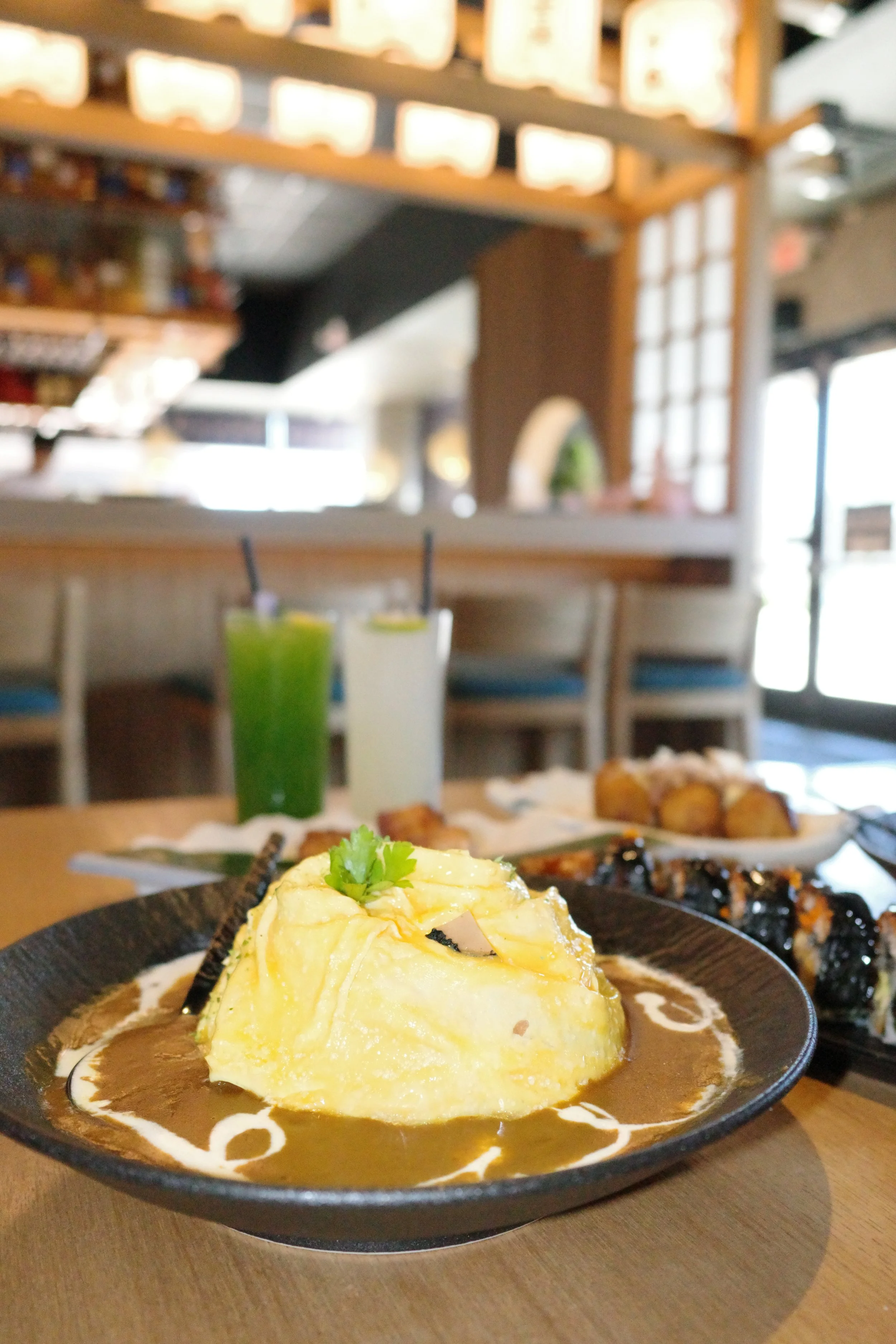 A plate with a large serving of mashed potatoes topped with a sprig of cilantro, surrounded by brown gravy with white decorative streaks, on a wooden table. In the background, two drinks, possibly green juice and a creamy beverage, are visible along 