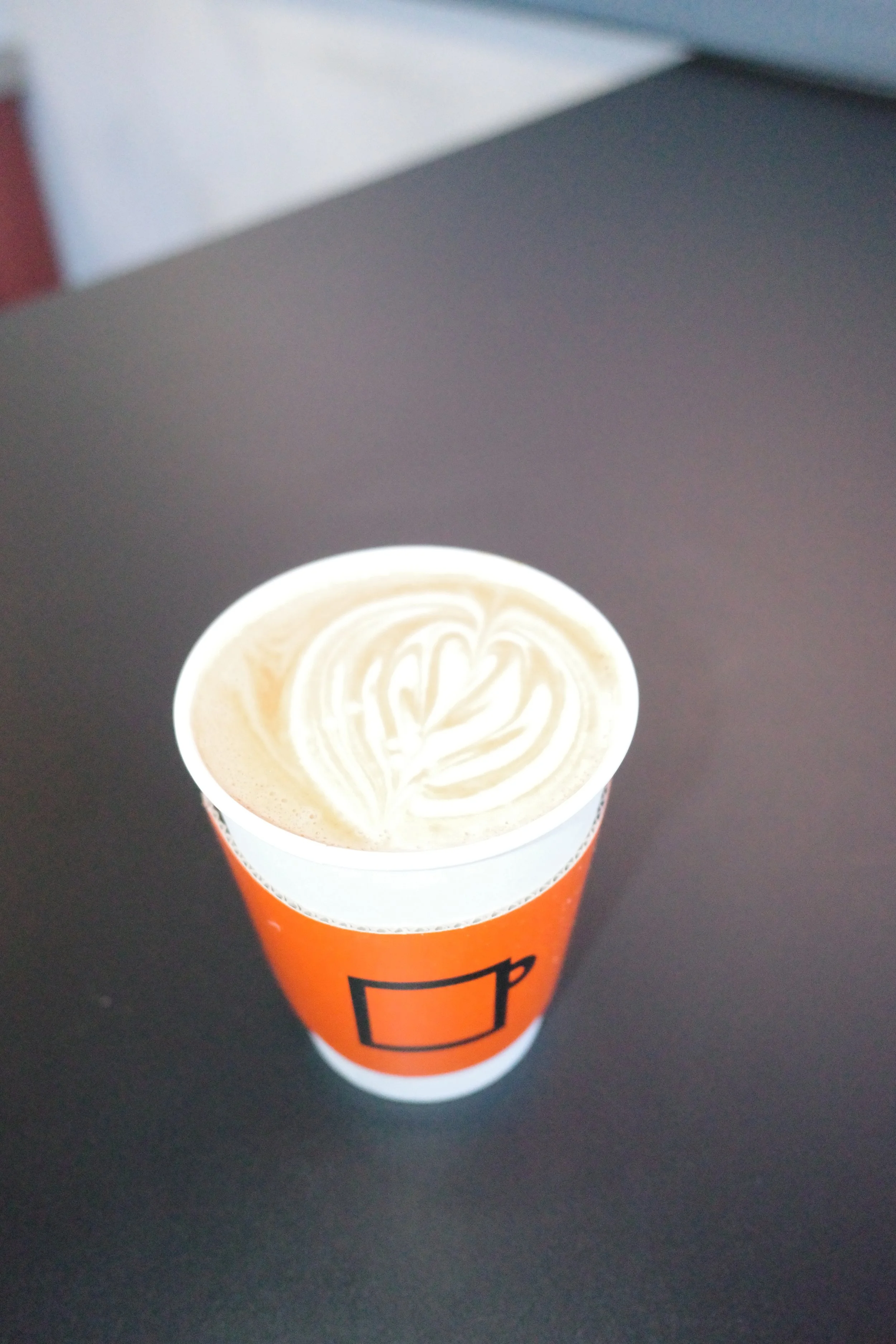 A cup of coffee with latte art, placed on a black surface, with a paper sleeve featuring a black outline of a mug on an orange background.
