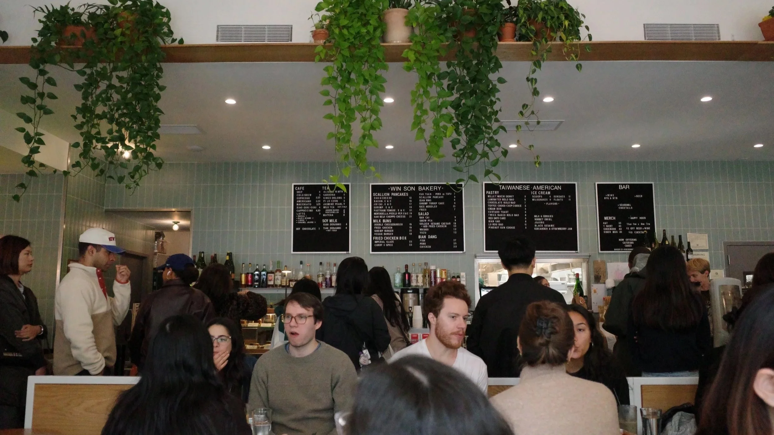 Crowded cafe with people waiting in line and sitting at tables, green hanging plants above with menu boards on the wall behind the bar area.