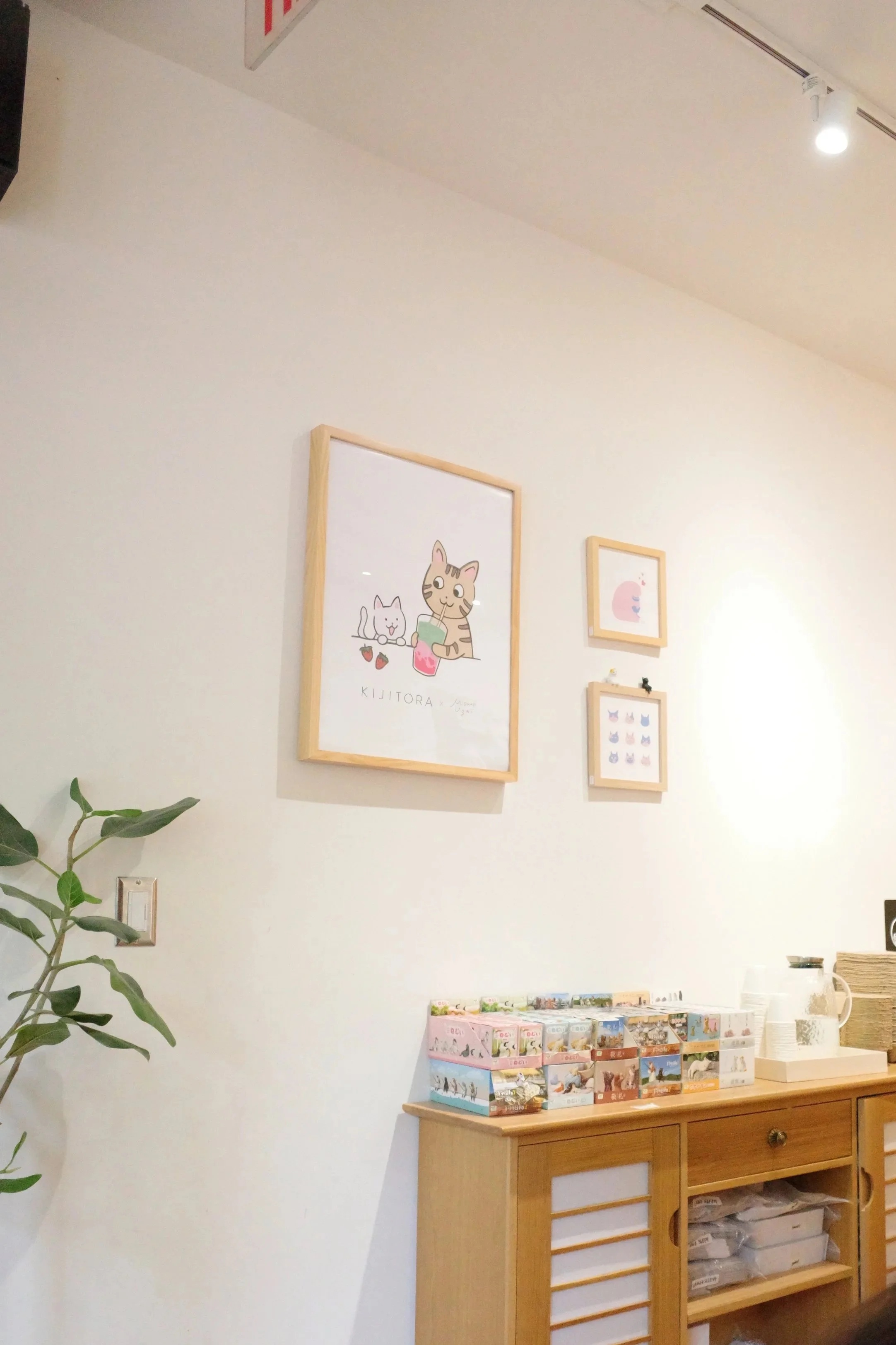 A corner of a room with a wooden cabinet on the right side, displaying colorful postcards or small posters, and framed pictures on the wall above. A green leafy plant is visible on the left side, and a white light fixture is on the ceiling. The pictu