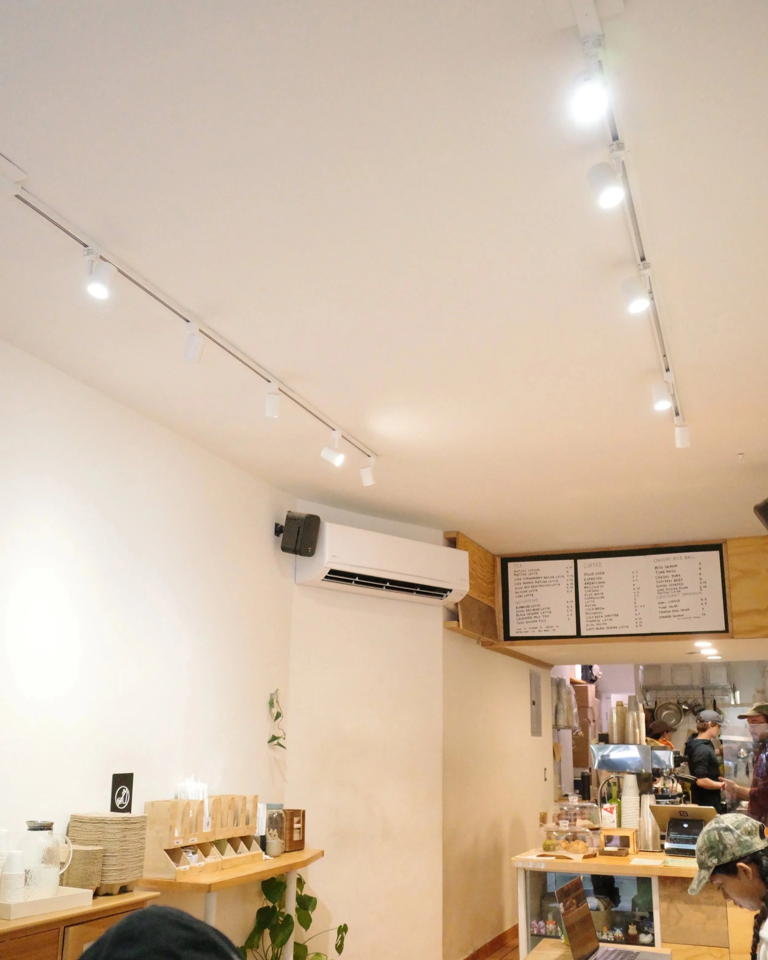 Interior of a cafe with a menu board on the wall, a counter with coffee equipment, and customers. The ceiling has spotlights, and there are some small items on a table.