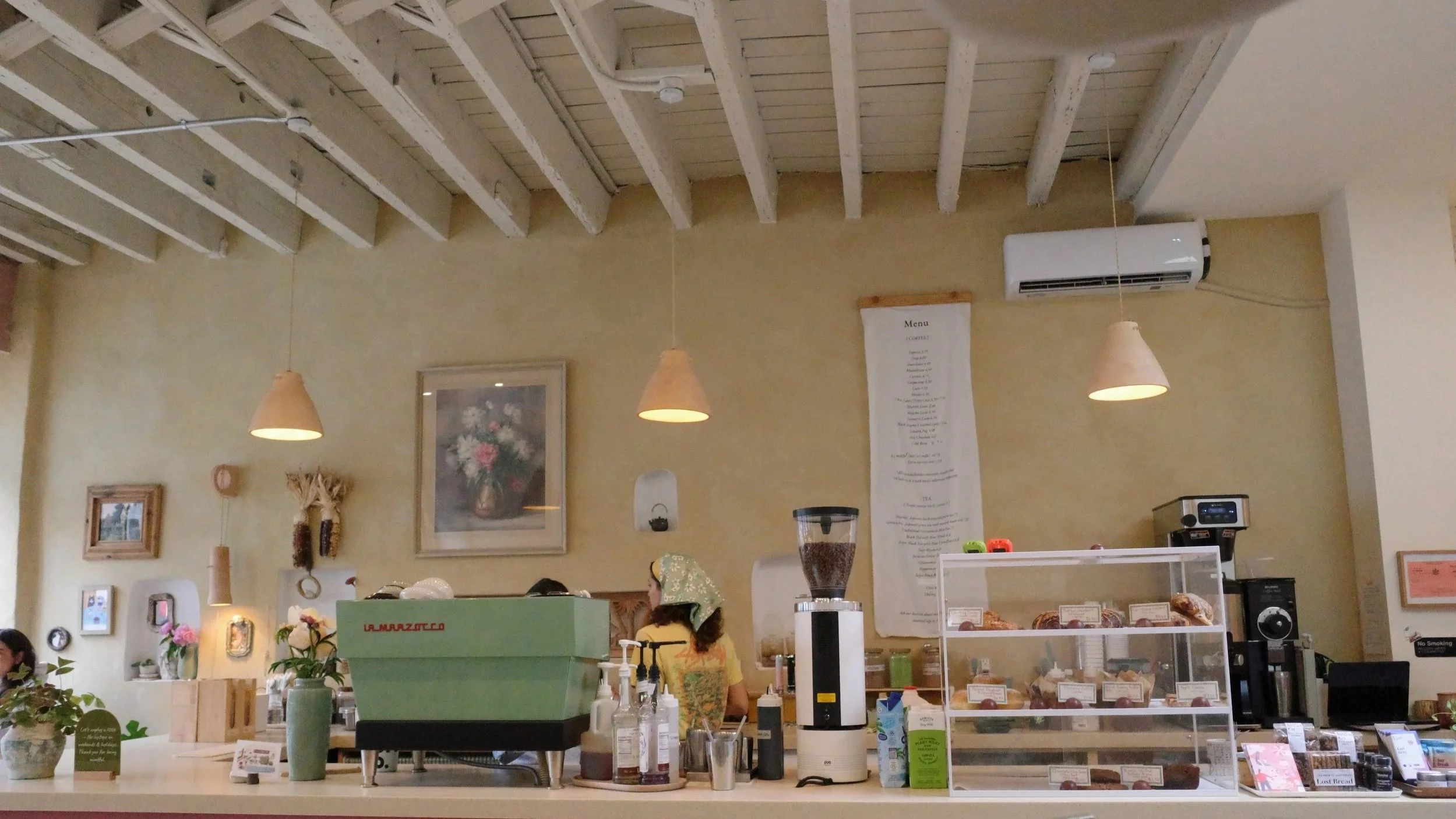 Inside a cozy cafe with light yellow walls, wooden ceiling beams, hanging pendant lights, framed art, flowers, a menu board, and a counter with baked goods and coffee equipment. Customers and barista are present.