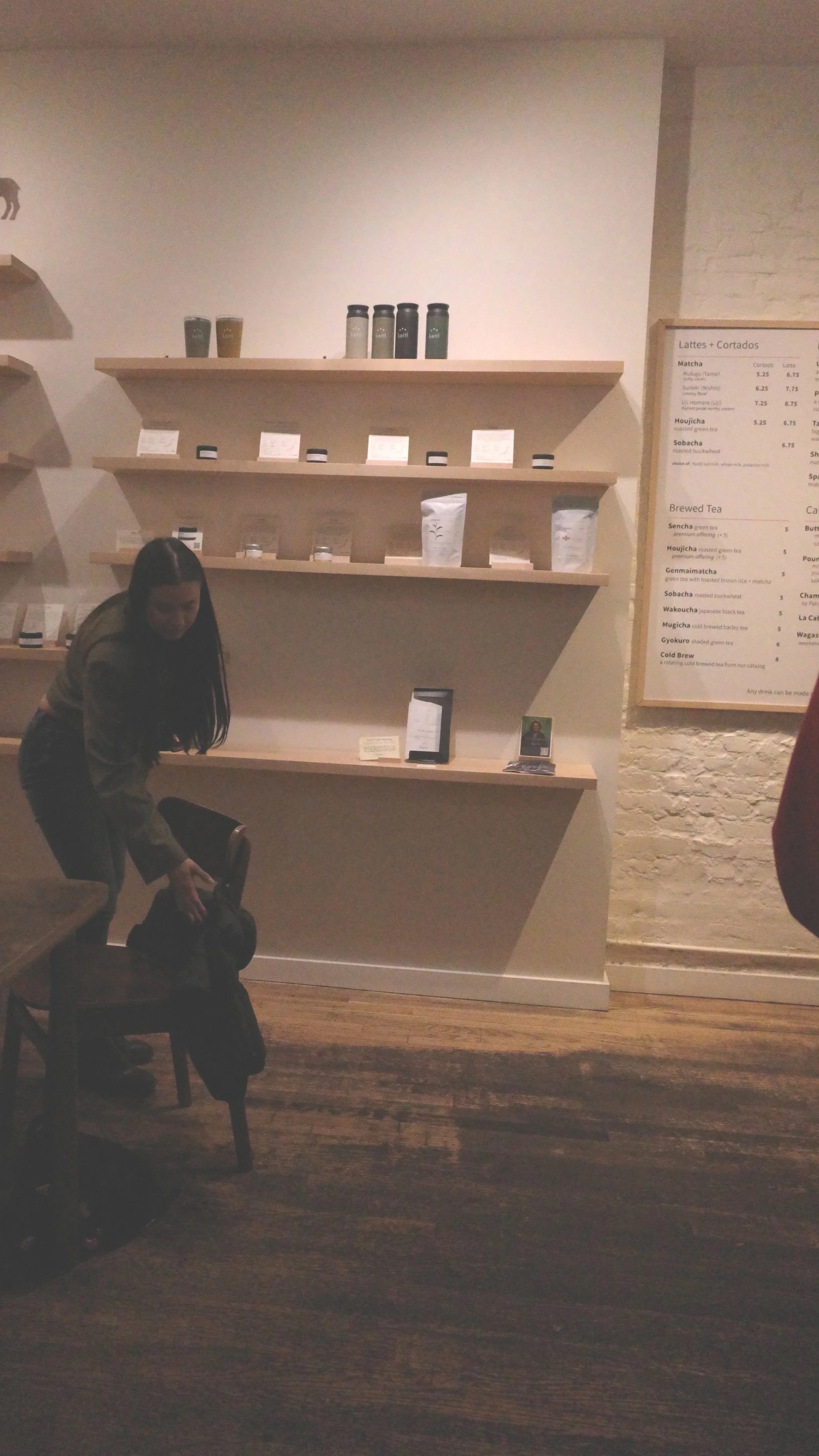 A woman reaching into a backpack on a chair in a coffee shop with shelves of coffee products and a menu board on the wall.