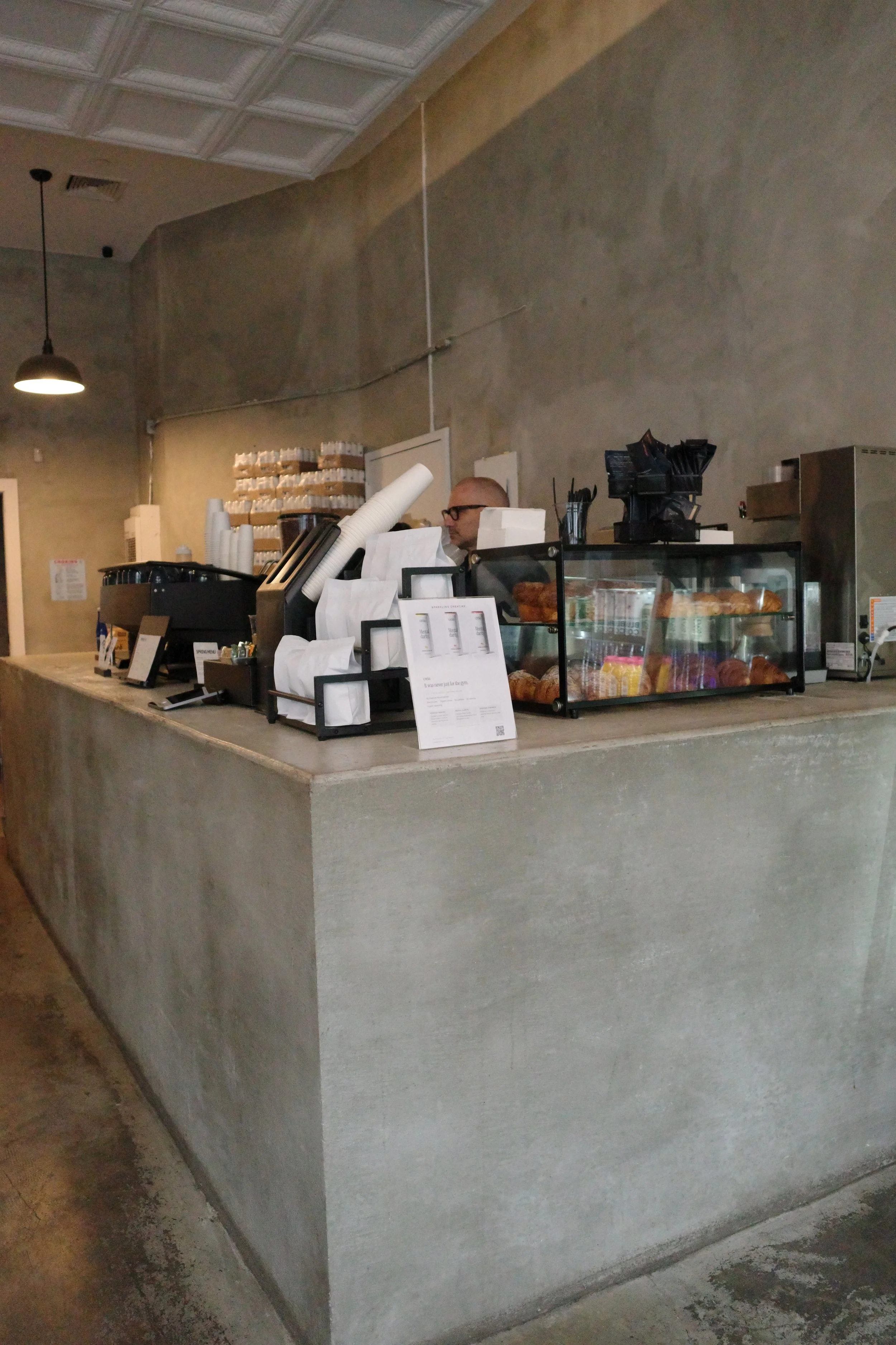 View of a coffee shop counter with coffee brewing equipment, pastries on display, and a barista behind the counter, in a modern minimalist style with concrete walls and ceiling.