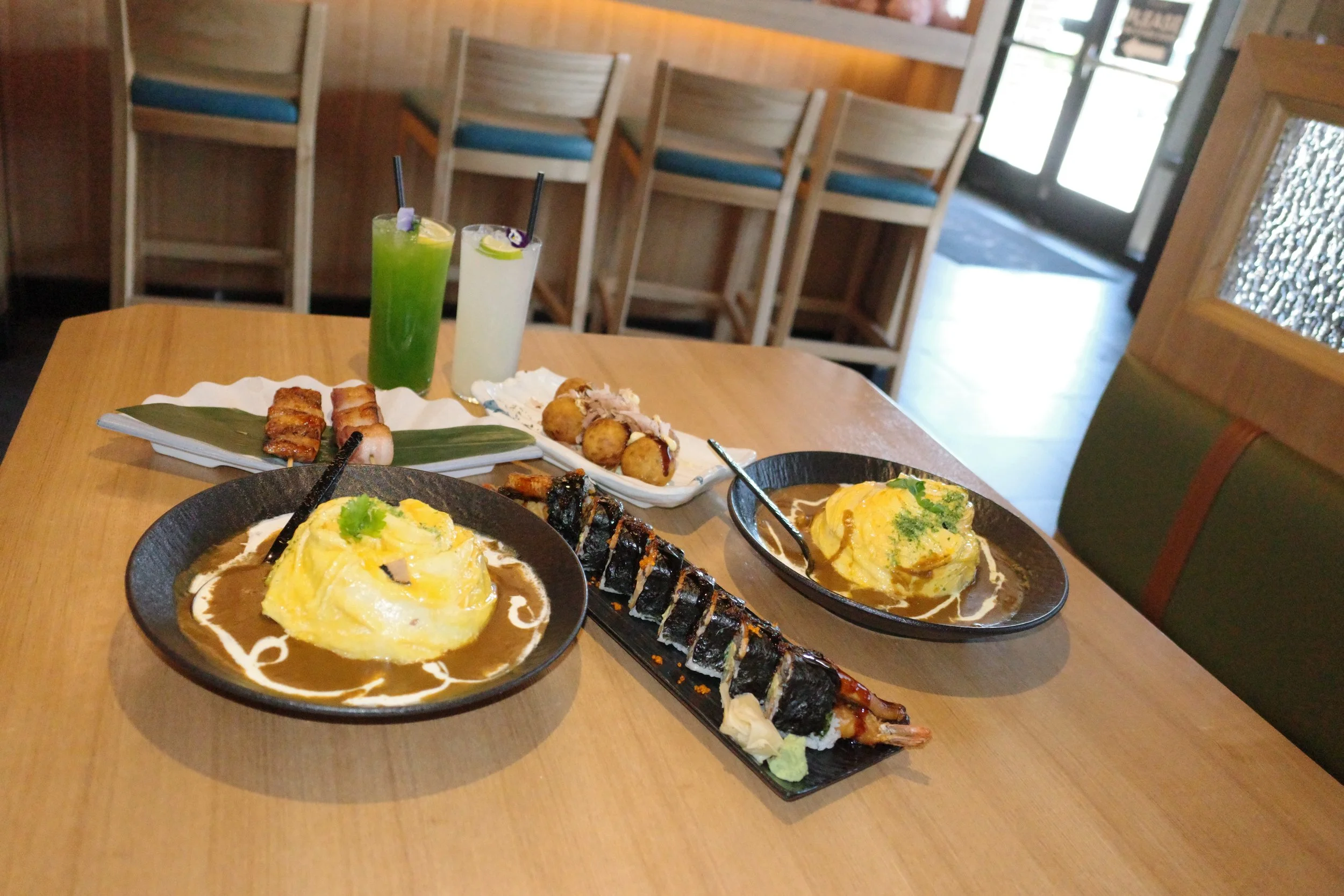 A wooden table with Japanese food, including two bowls of omurice topped with sauce, a plate of grilled eel sushi, a plate of skewered grilled meat and vegetables, and two colorful cocktails in tall glasses, in a restaurant with wooden chairs and a g