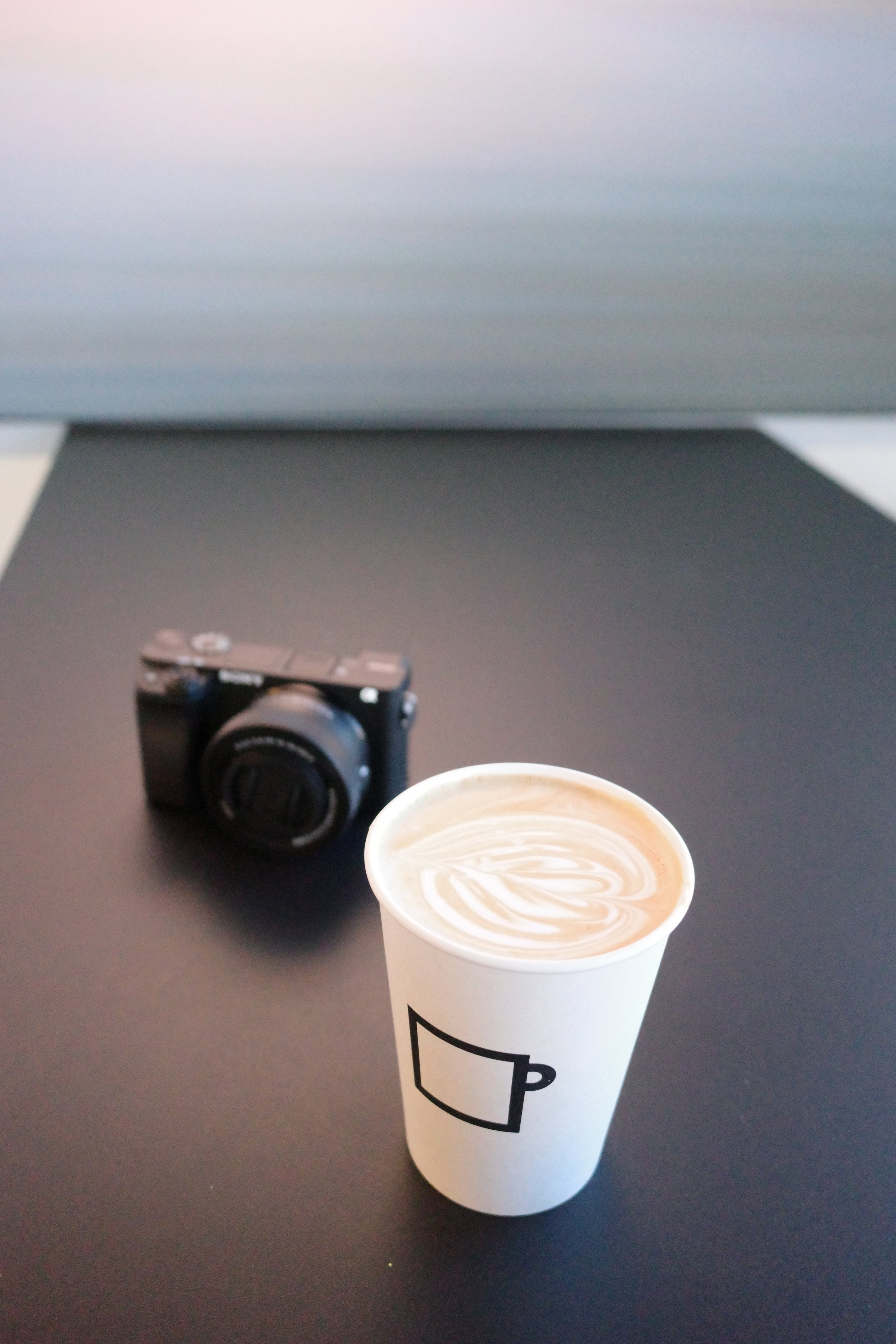 A paper cup with a coffee beverage featuring latte art, placed on a dark tabletop with a black camera and a laptop in the background.