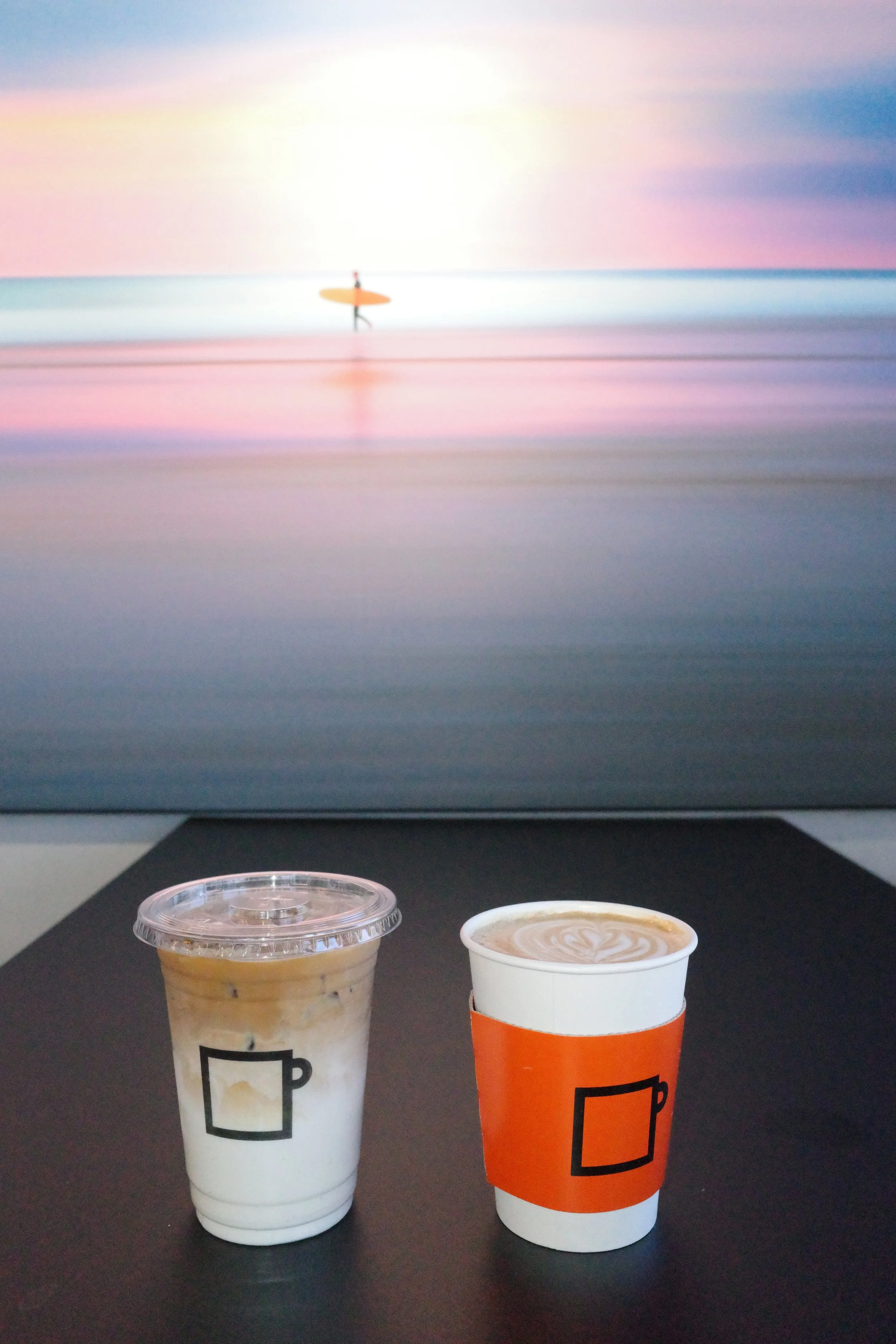 A computer screen displaying a beach scene with pastel colors and a person with a paddleboard in the water, positioned on a desk with two coffee cups in front of it.
