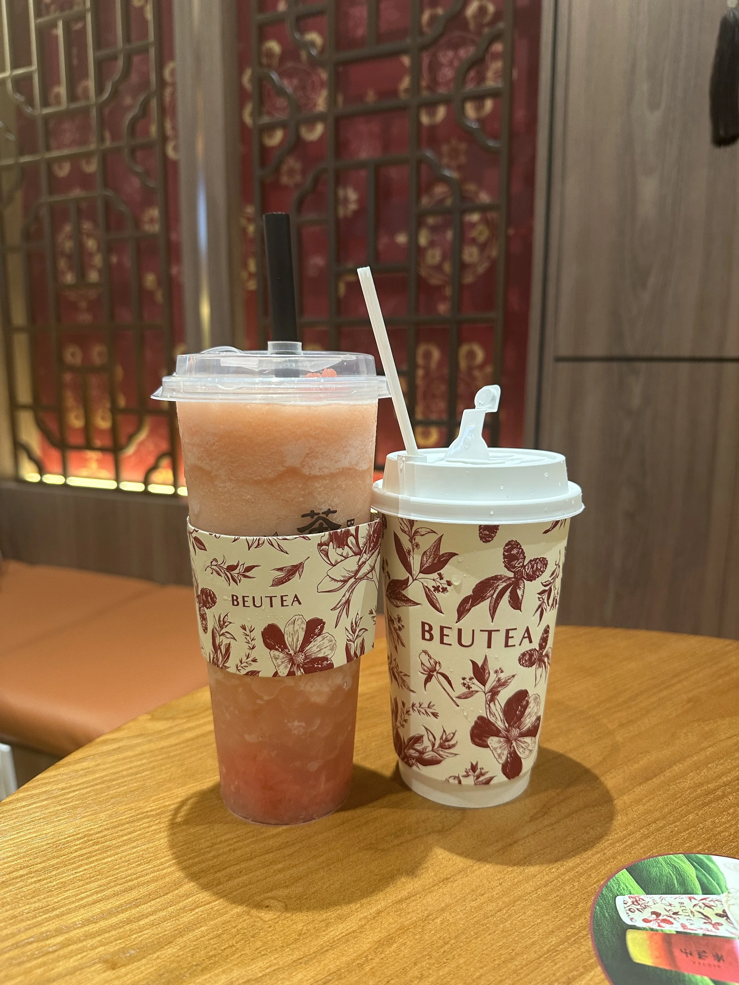 Two drinks in disposable cups with floral designs and the word 'BEUTEA' on them, placed on a wooden table in a restaurant or cafe setting.