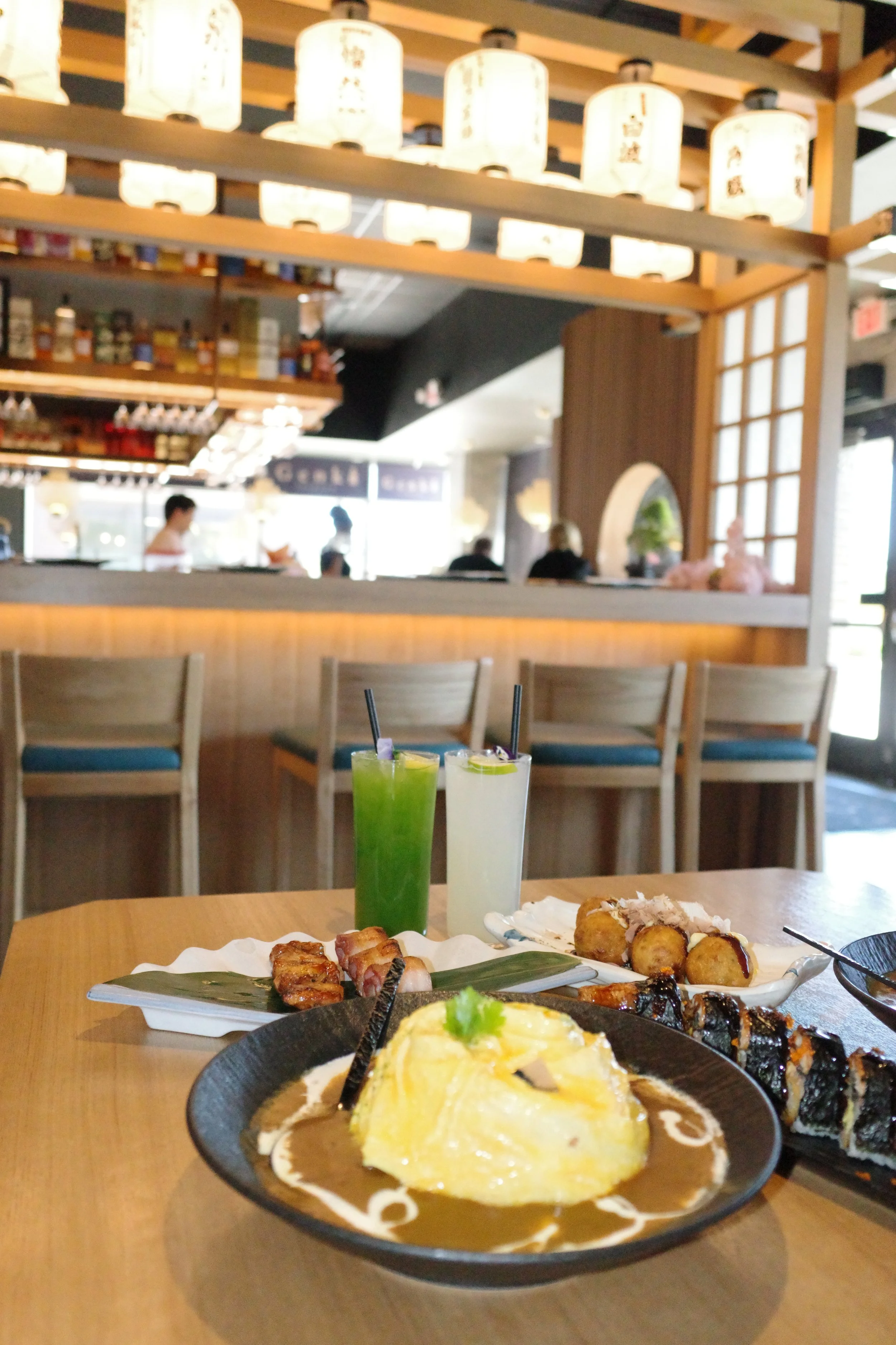 A restaurant table with Japanese food dishes, two cocktails, and a background of a Japanese-themed interior with hanging lanterns and seated diners.