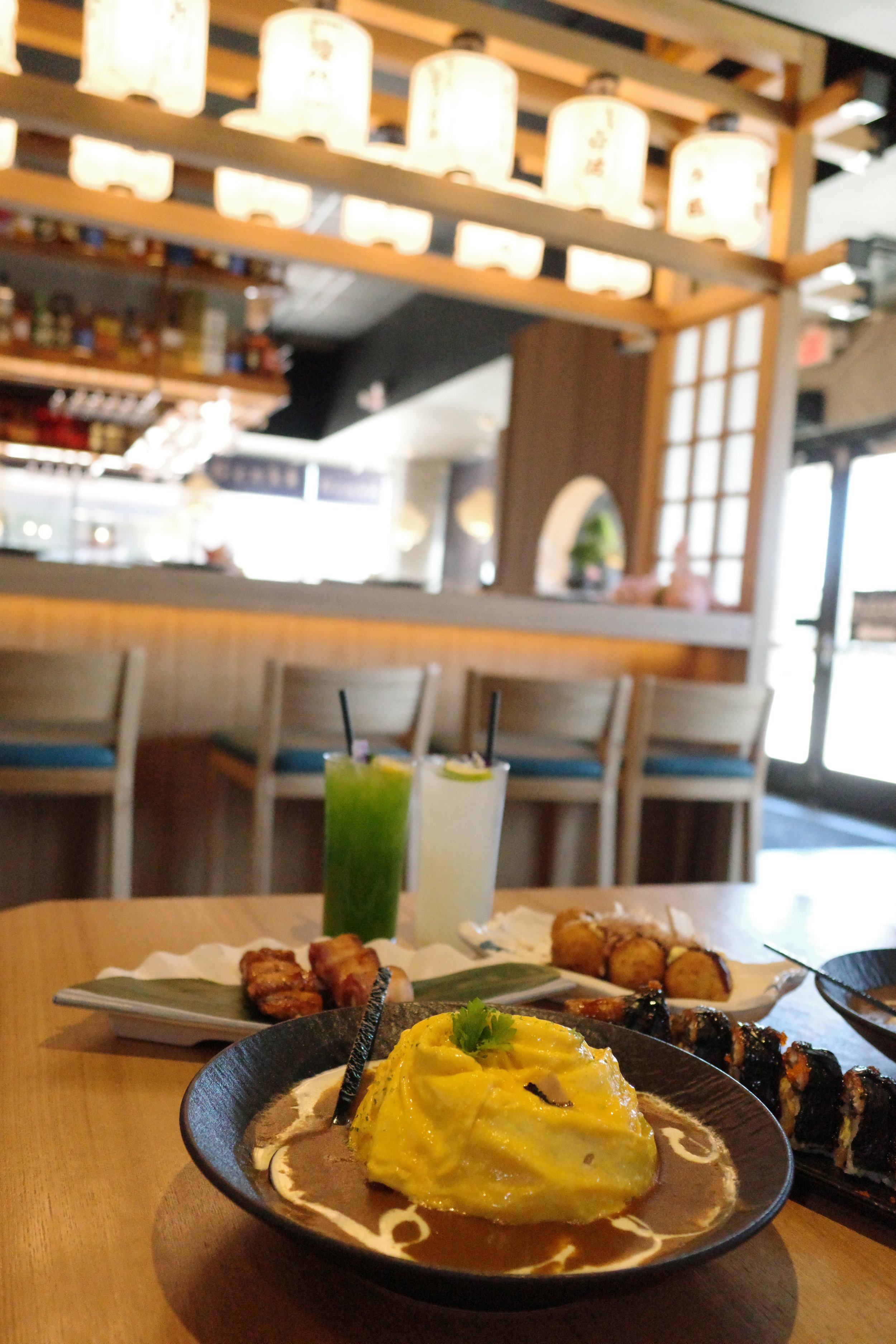 Plate of Japanese dishes including a yellow omelette with garnish, grilled meat skewers, and fried items, with drinks on a wooden table in a restaurant with warm lighting and lantern decor.