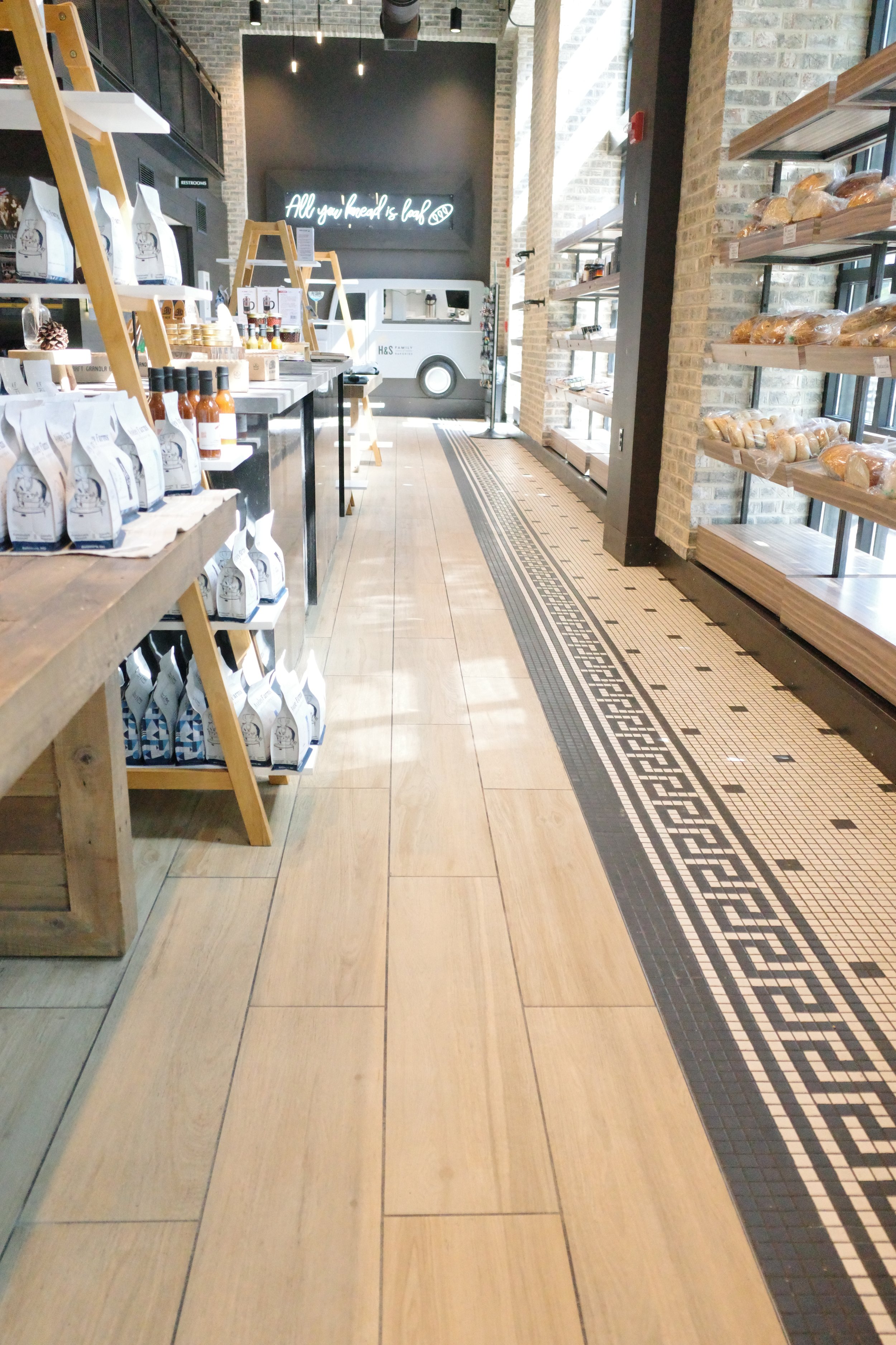 Interior of a modern bakery with wooden shelves displaying various baked goods and bottled sauces, tiled flooring with a pattern, brick walls, a neon sign that reads 'All you need is love,' and large windows letting in natural light.