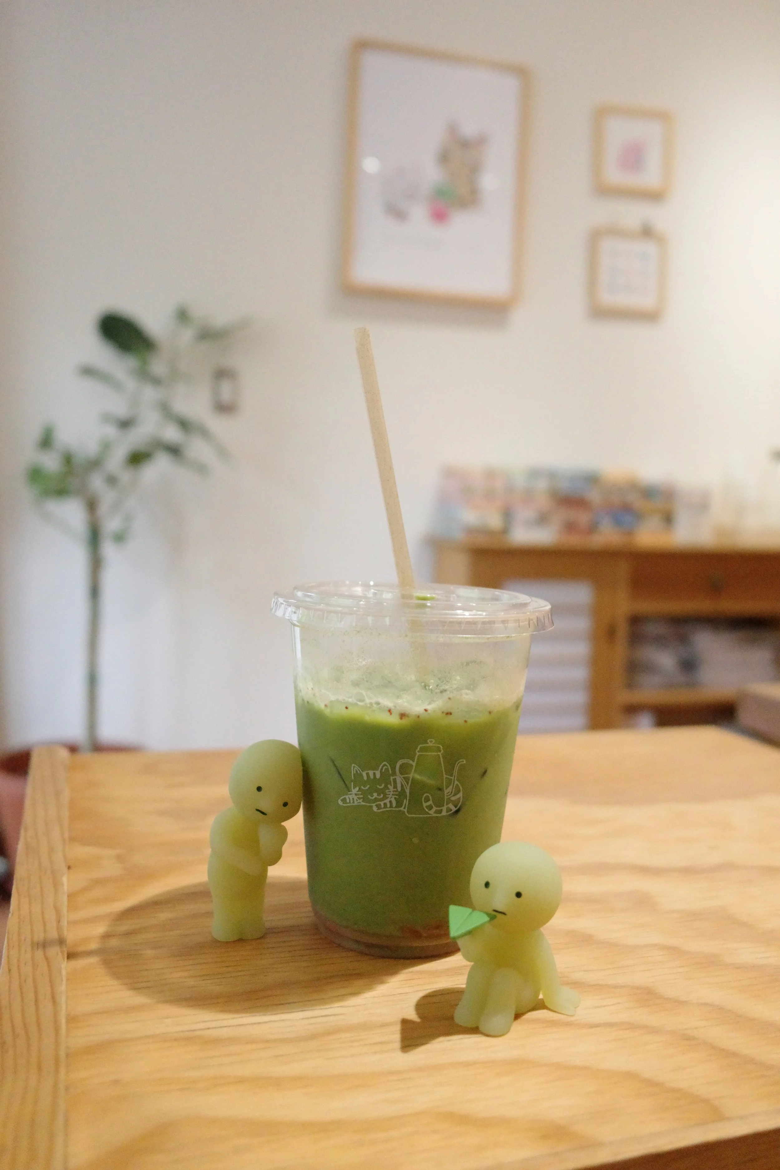 A green matcha latte in a clear plastic cup with a brown straw, surrounded by small beige bunny figurines on a wooden tray, with a blurred background of a beige wall with framed pictures and a green plant.