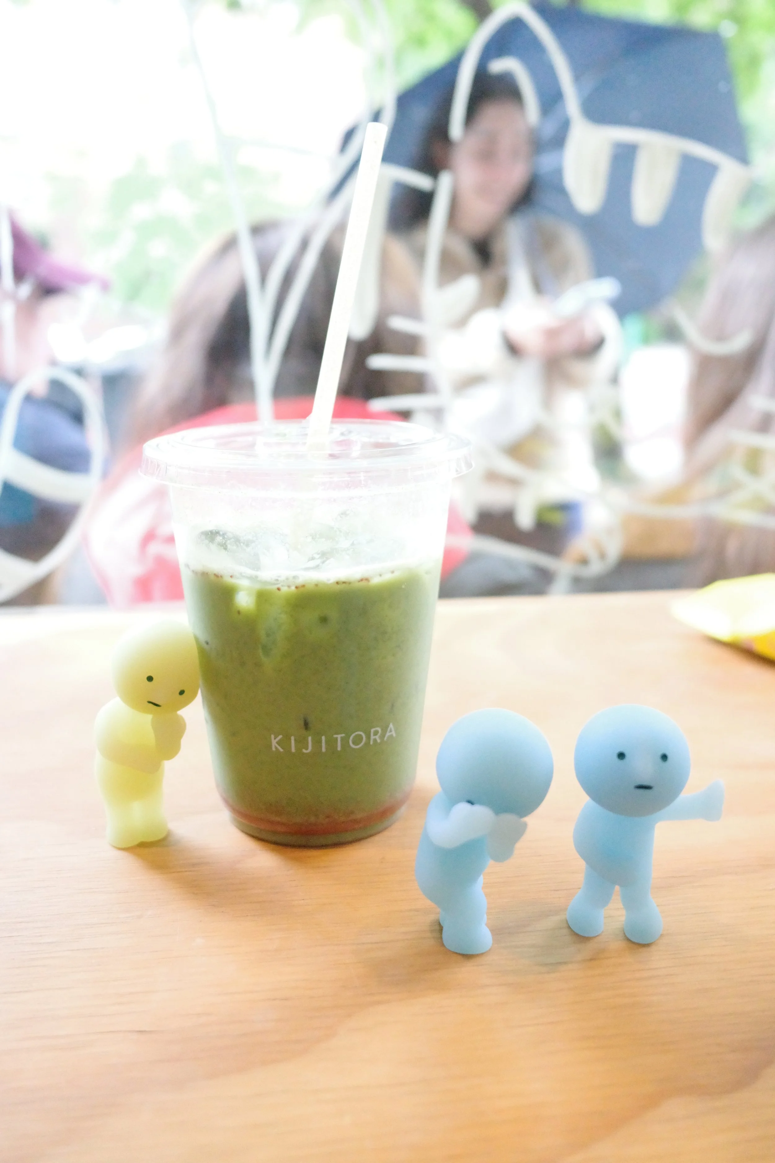 A cup of green smoothie from KIJIITORA on a wooden table, surrounded by small yellow and blue figurines with cute, simple faces.