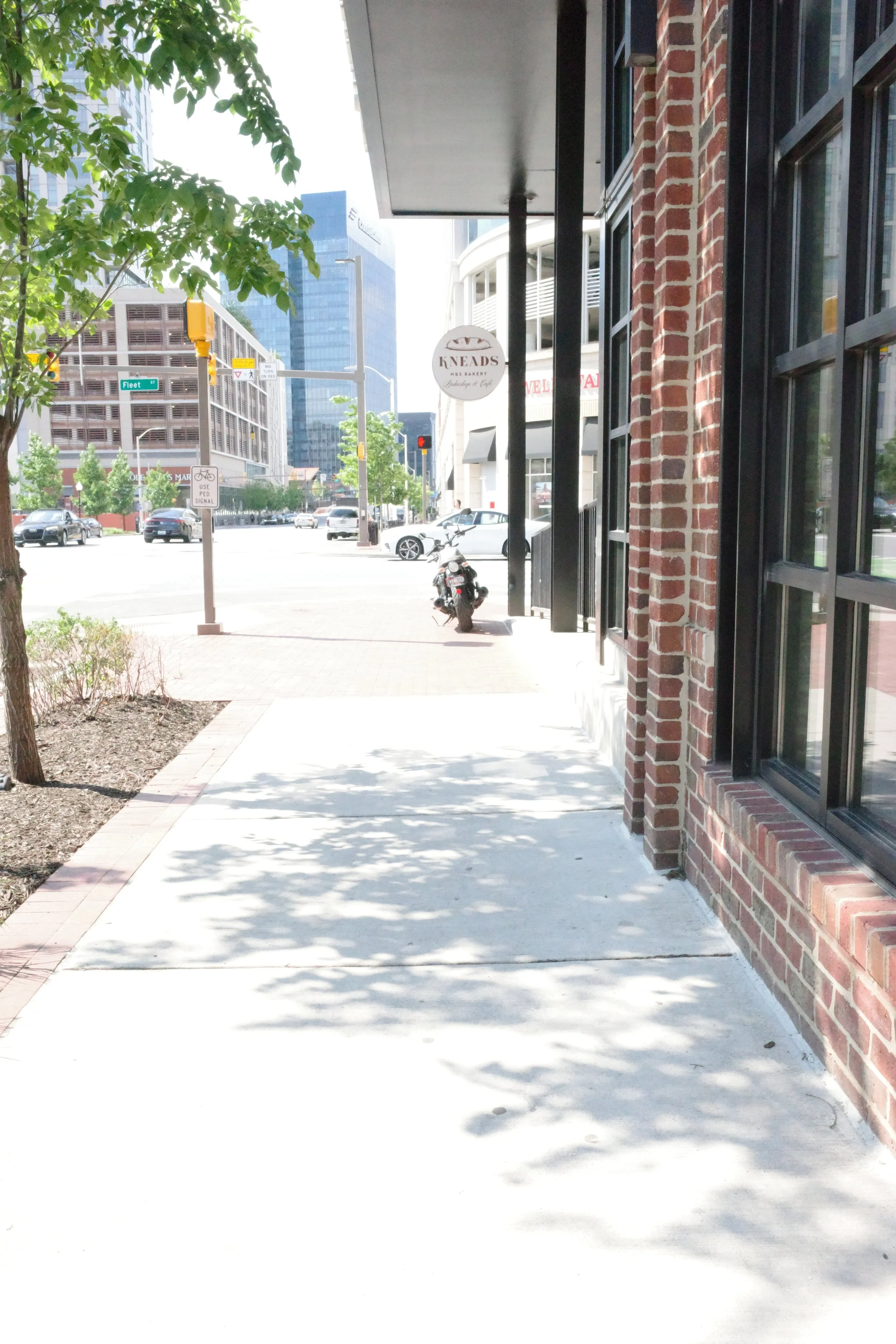 Street scene with a sidewalk, brick building, pedestrians, parked motorcycles, cars, and modern glass high-rise buildings in the background.