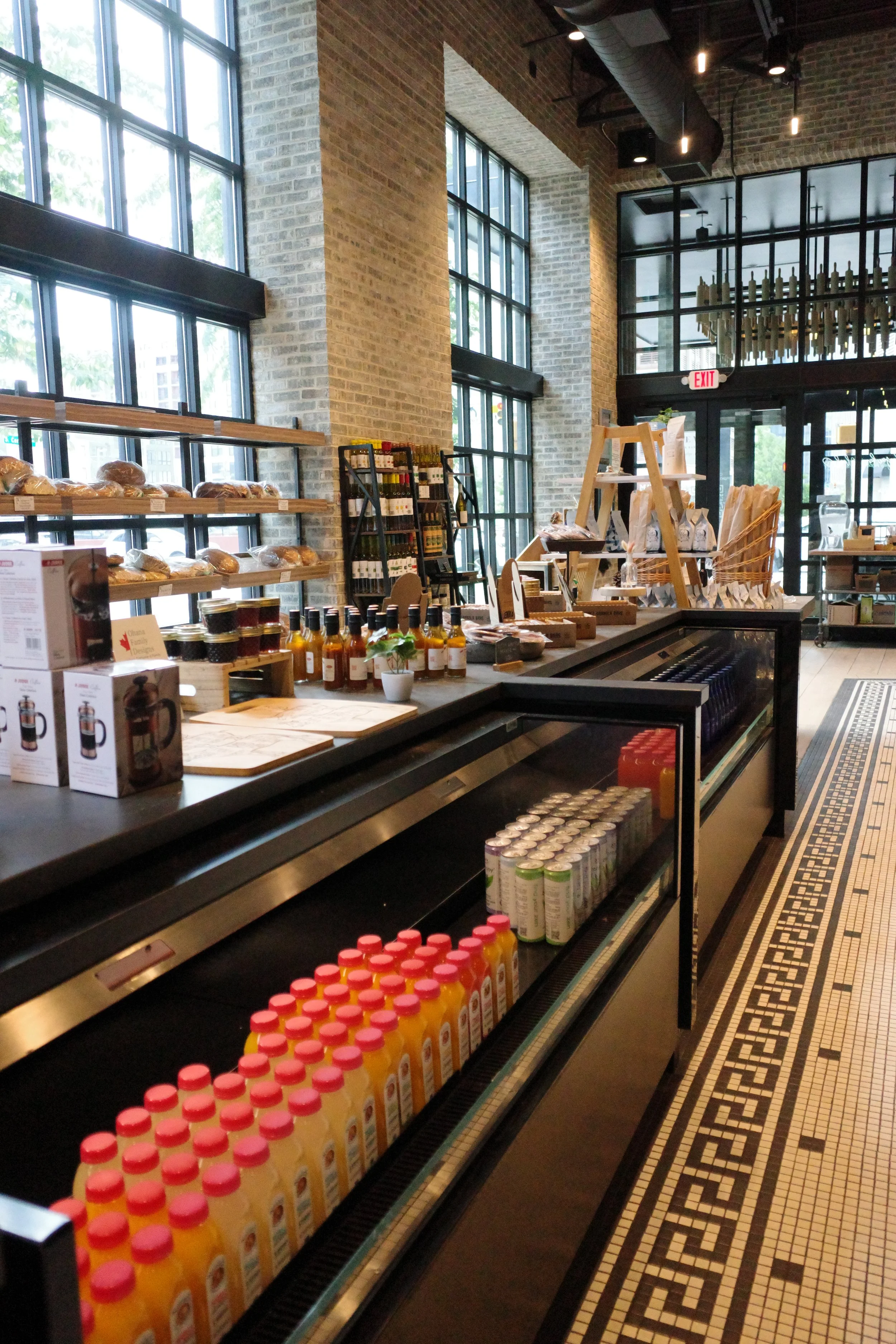 Interior of a grocery or convenience store with shelves of bread, bottles of sauces, and juice bottles, large windows, brick walls, and a black and white patterned tiled floor.