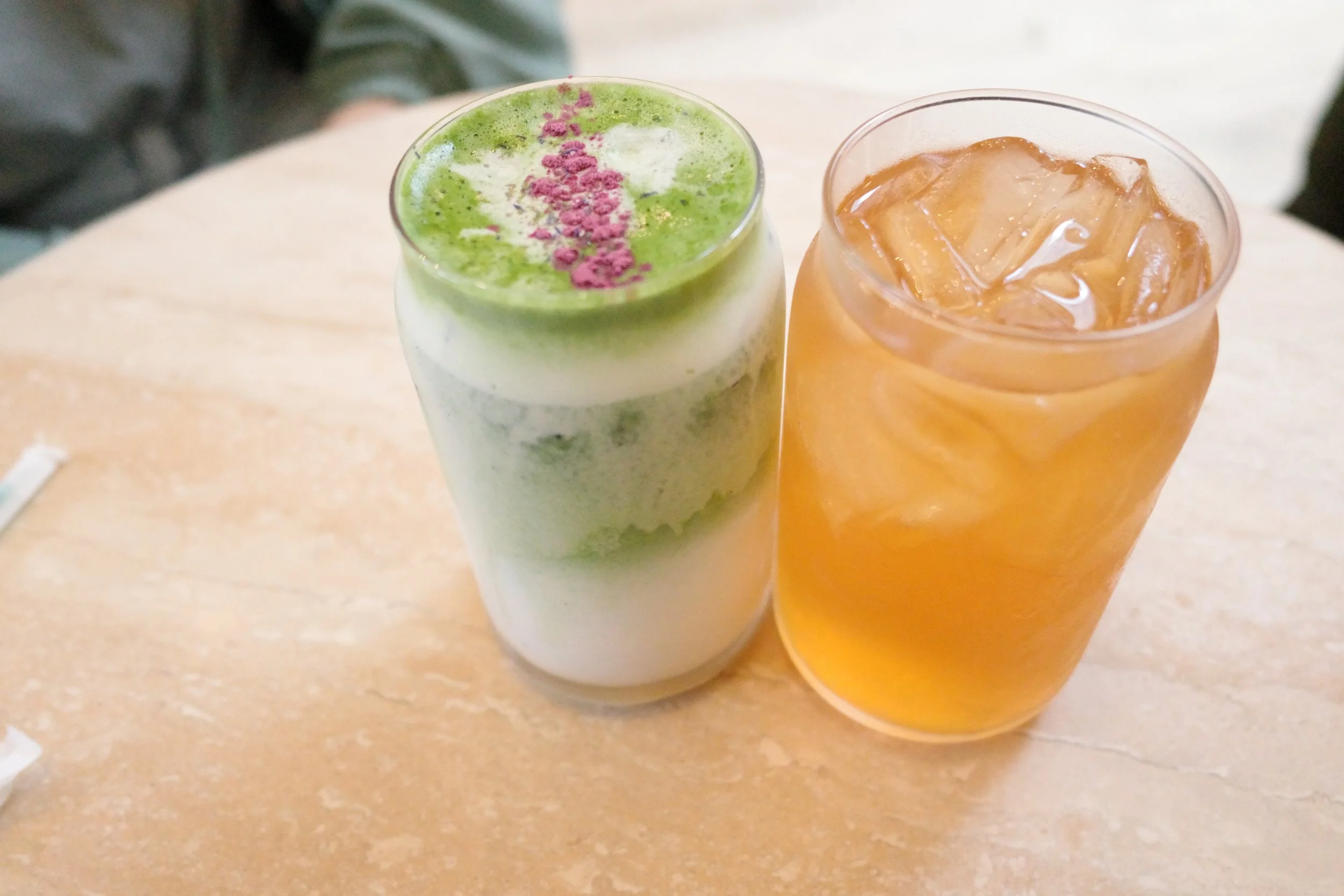 Two drinks on a beige table, one is a layered green and white beverage topped with pink crumbs, the other is a light amber iced drink.
