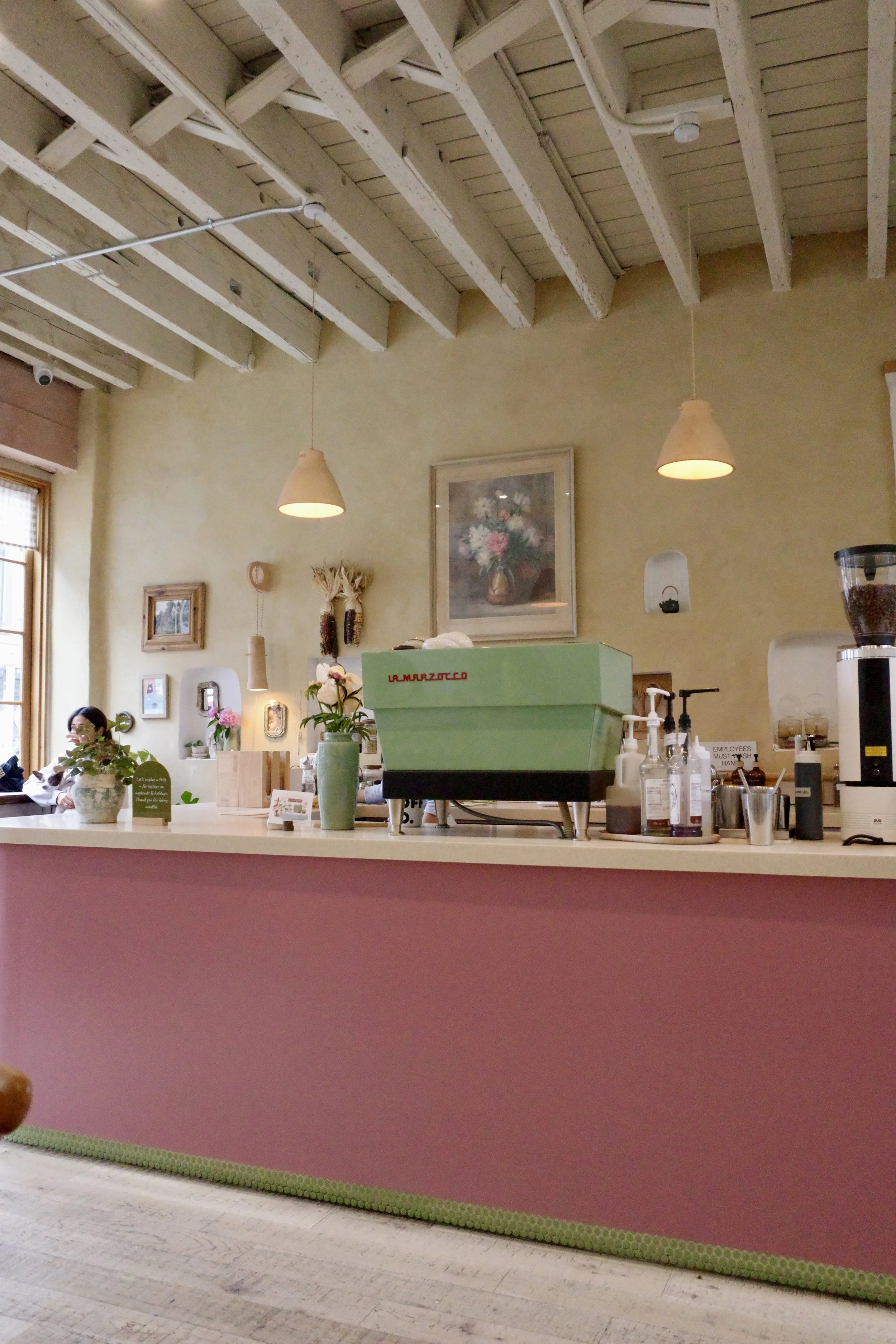 Interior of a cafe or coffee shop with a pink counter, decorative plants, framed pictures, hanging pendant lights, and a wooden ceiling with white beams.