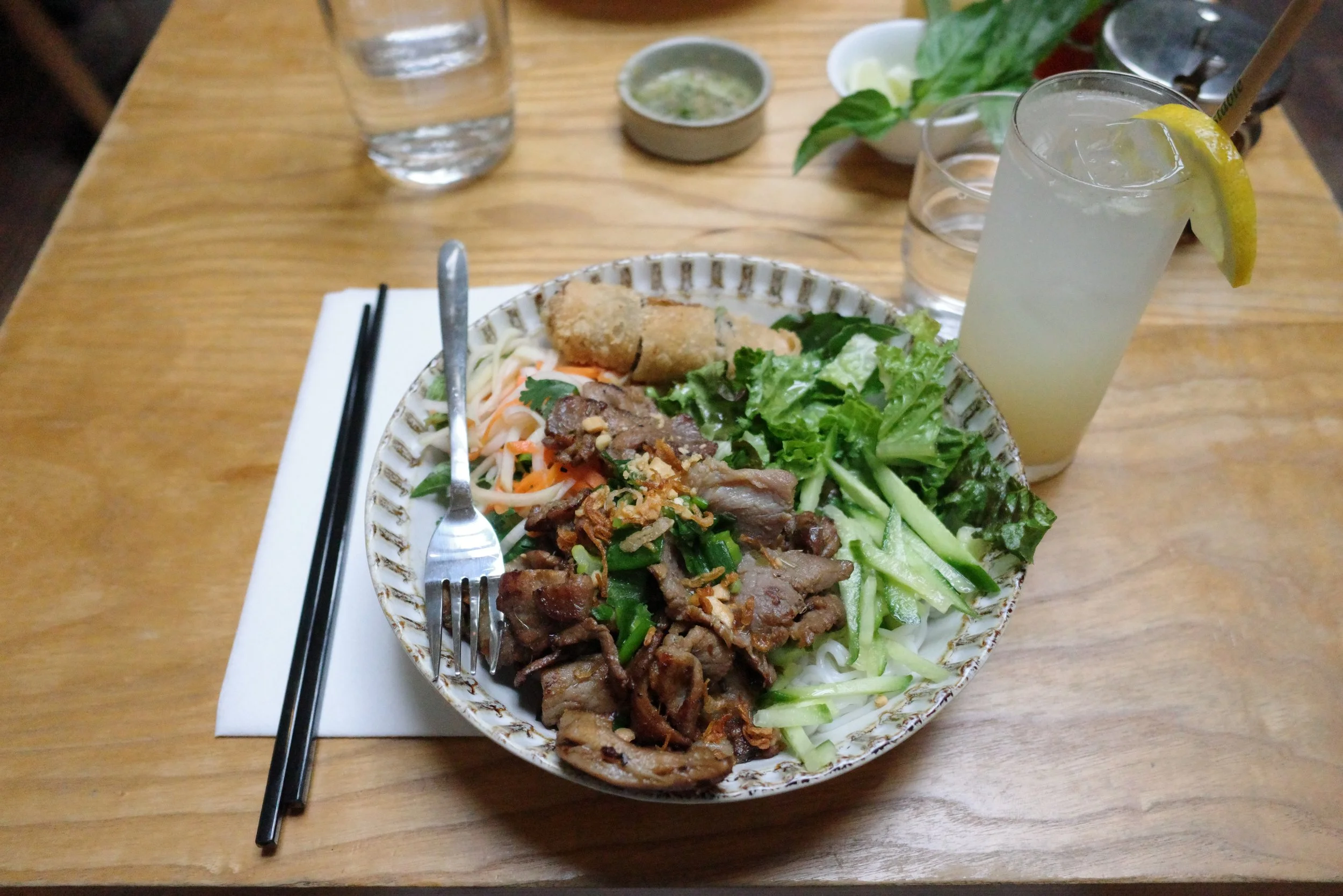 A plate of Vietnamese food with sliced beef, fried spring roll, fresh herbs and vegetables, with chopsticks and a fork on the side, on a wooden table. A glass of lemonade with a lemon slice and straw is beside the plate, and there are glasses of wate