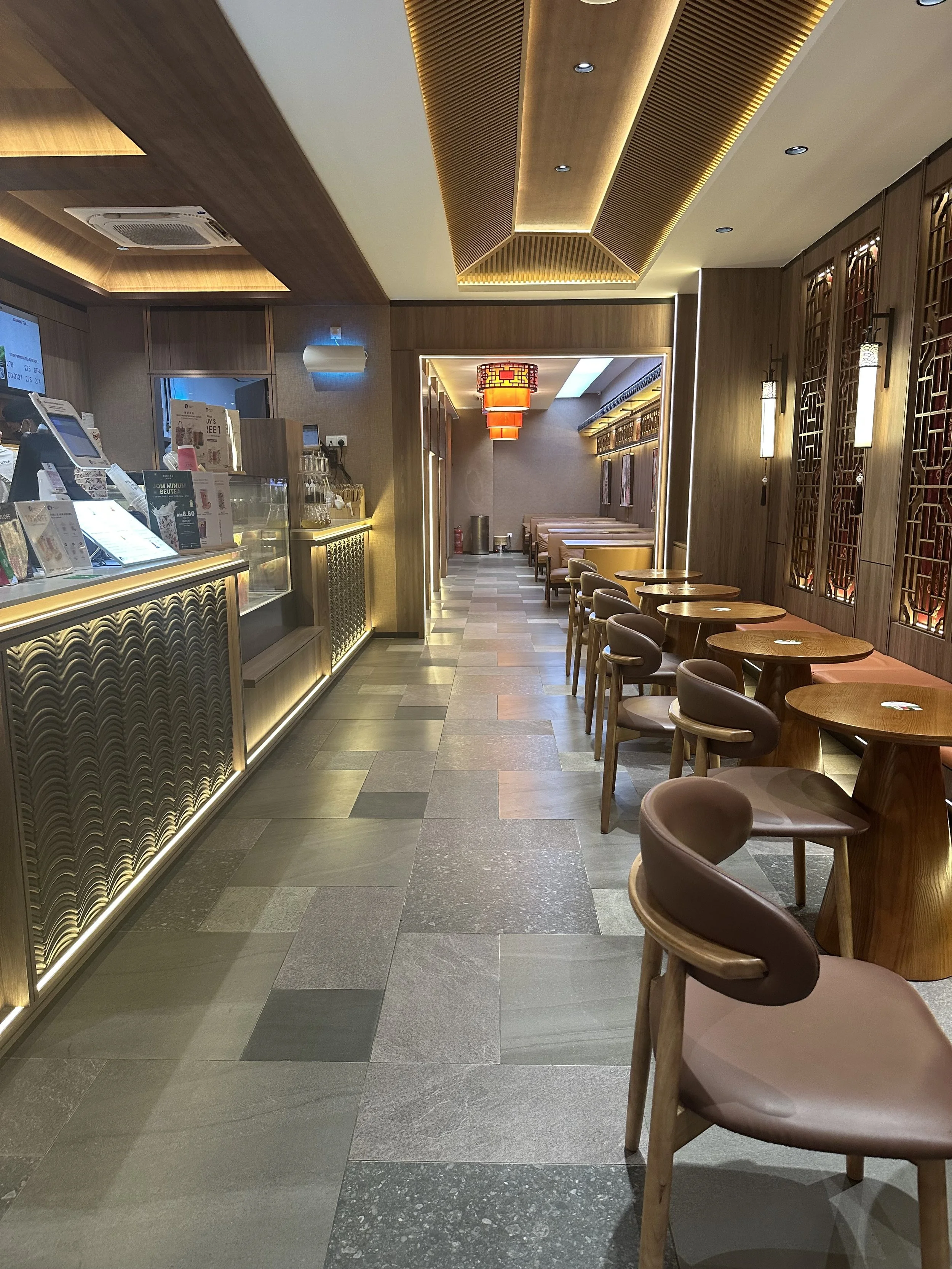 Interior of a modern Asian restaurant or café with wooden decor, round wooden tables, and beige chairs. The ceiling features wooden panels and warm lighting, with a red Chinese lantern hanging in the background. The floor is tiled with gray and brown