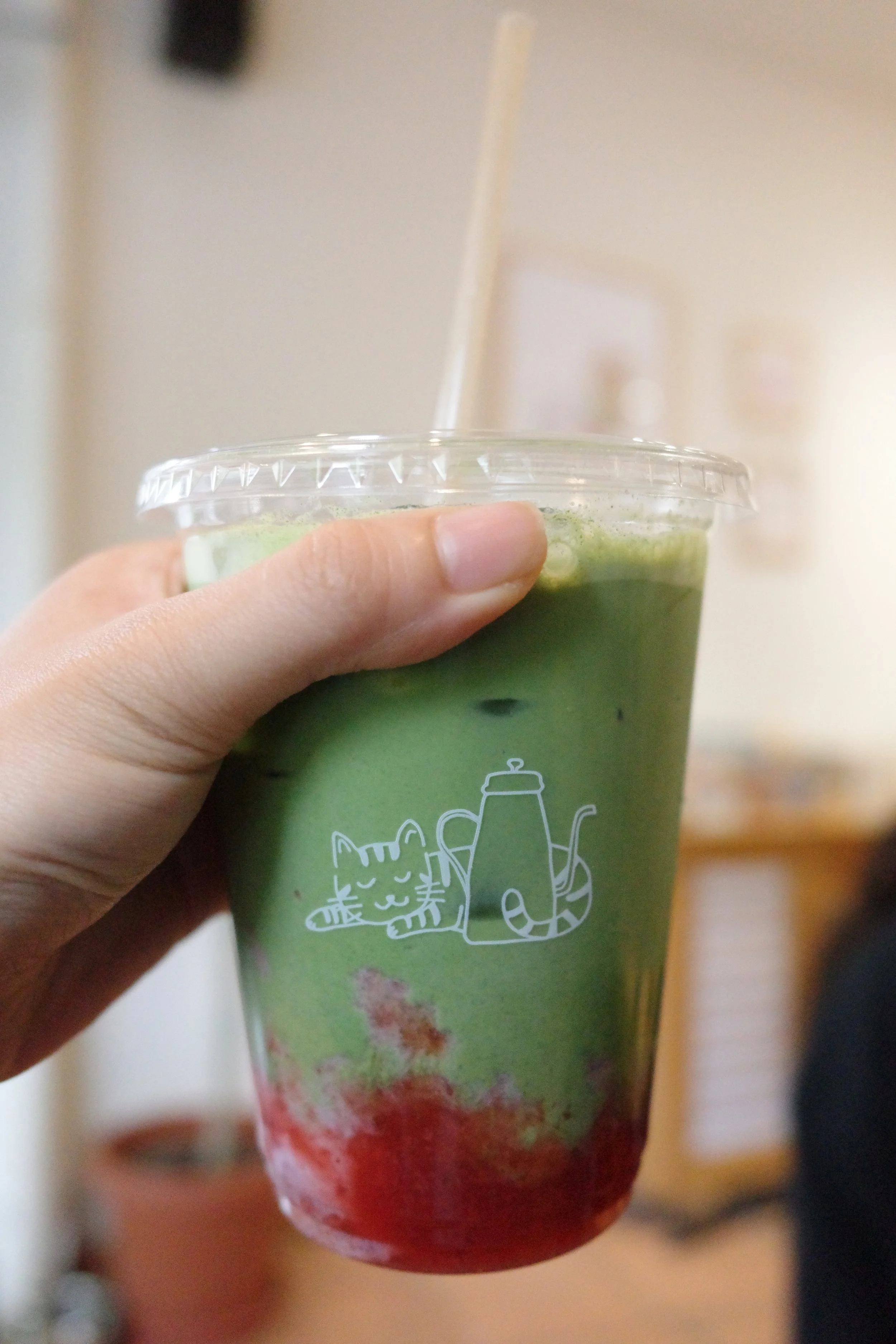 A hand holding a clear plastic cup filled with a layered green and red beverage. The cup has a white cartoon cat and plant illustration on it.