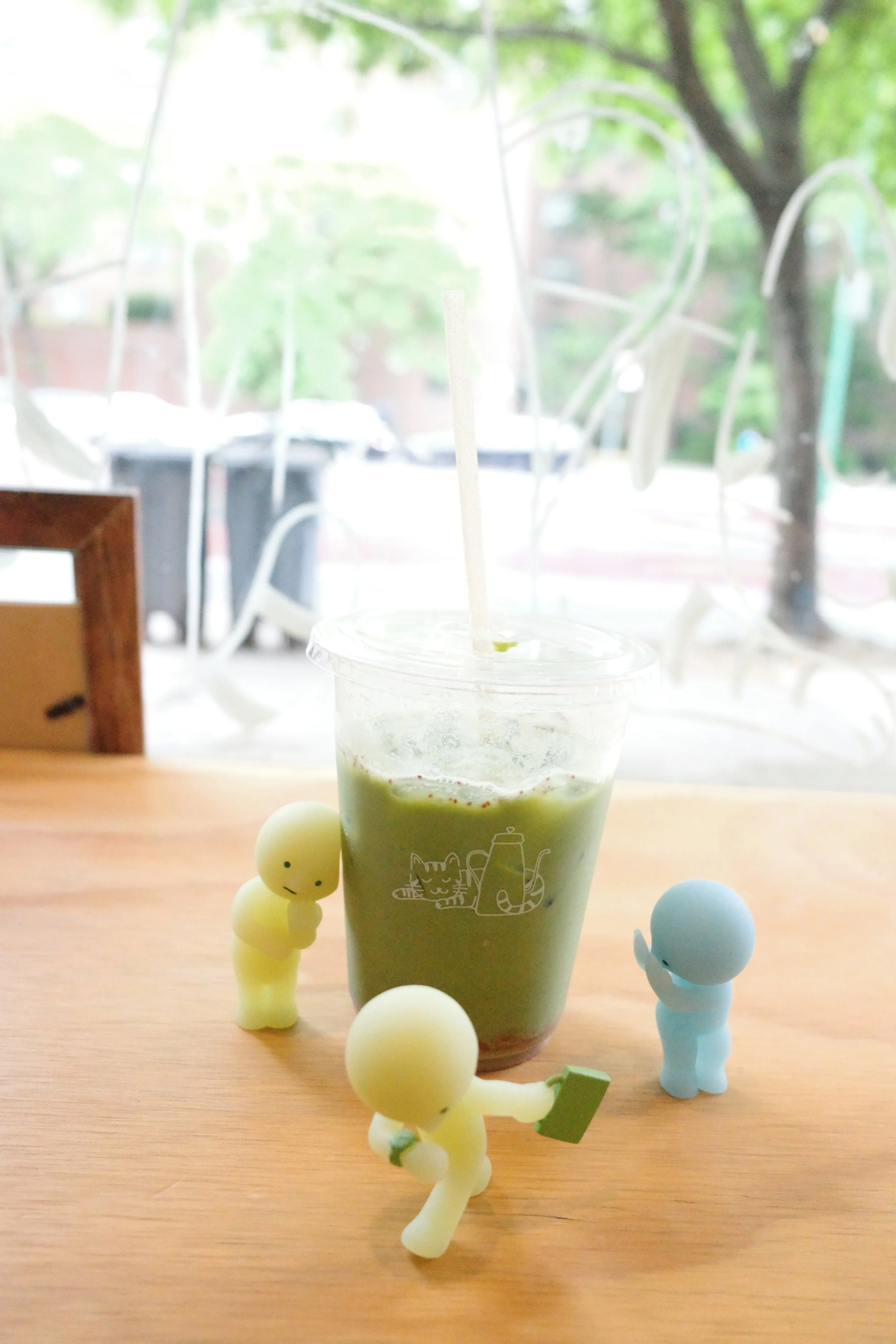 A green matcha drink in a clear cup with a straw, placed on a wooden table. Two small, cartoonish figurines resembling children or babies, one yellow and one blue, are standing nearby. The scene is set near a window with a blurred outdoor background 