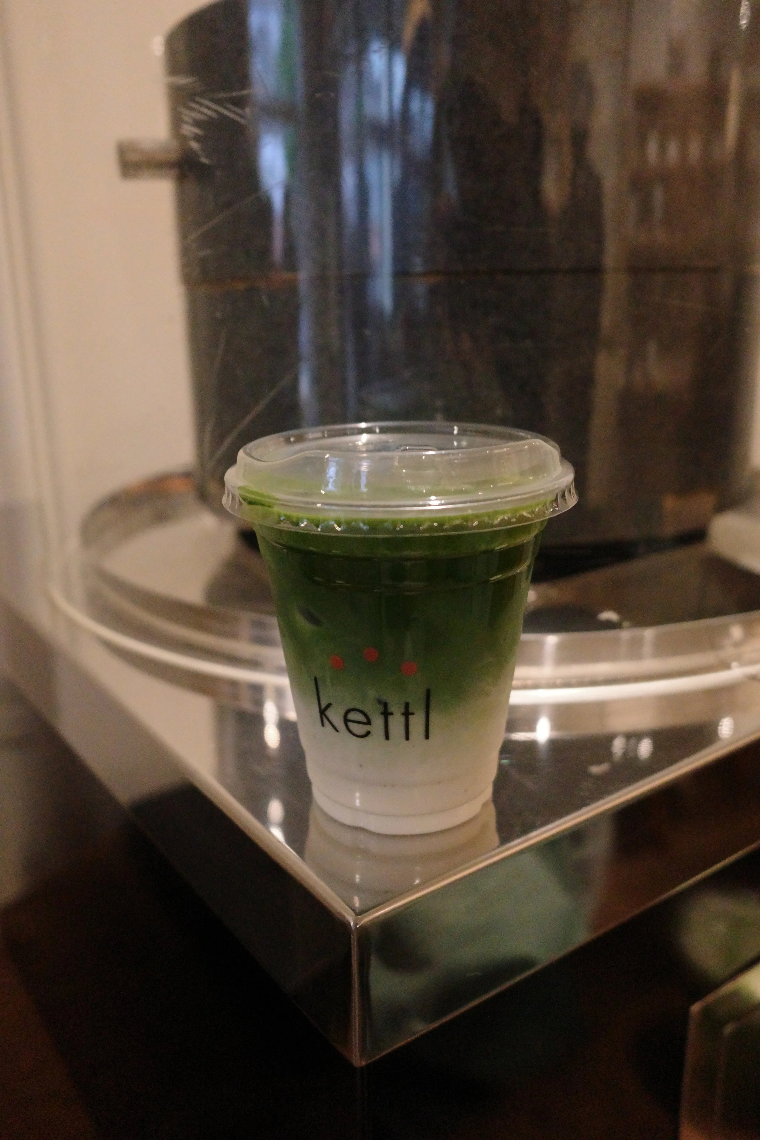 A plastic cup filled with a green beverage, positioned in front of a black coffee maker. The cup has the word 'Kettl' printed on it and is placed on a metallic tray.