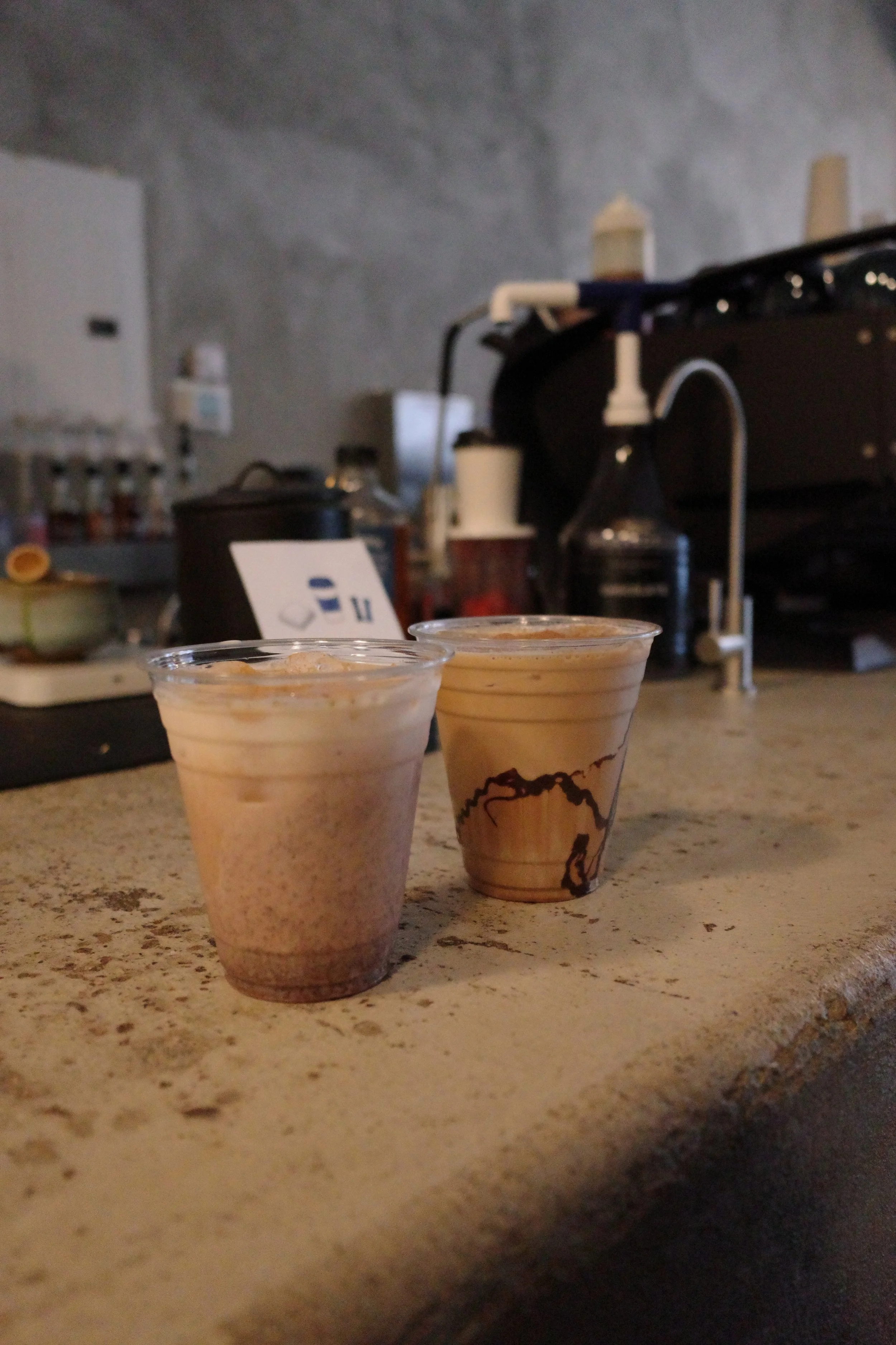 Two cups of iced coffee on a speckled beige countertop in a coffee shop setting, with a coffee machine and supplies in the background.