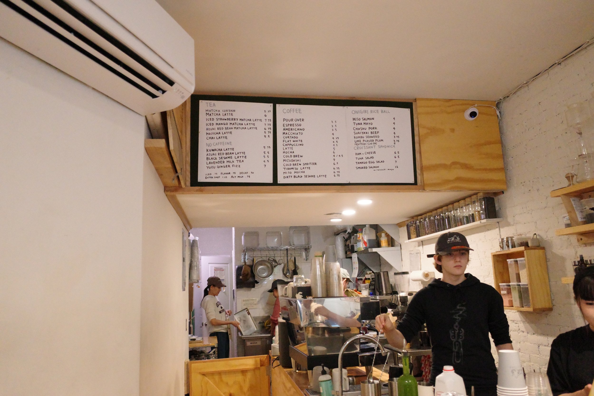 Inside a coffee shop with a menu board listing various drinks and rice bowl options. Staff members are working behind the counter, preparing orders.