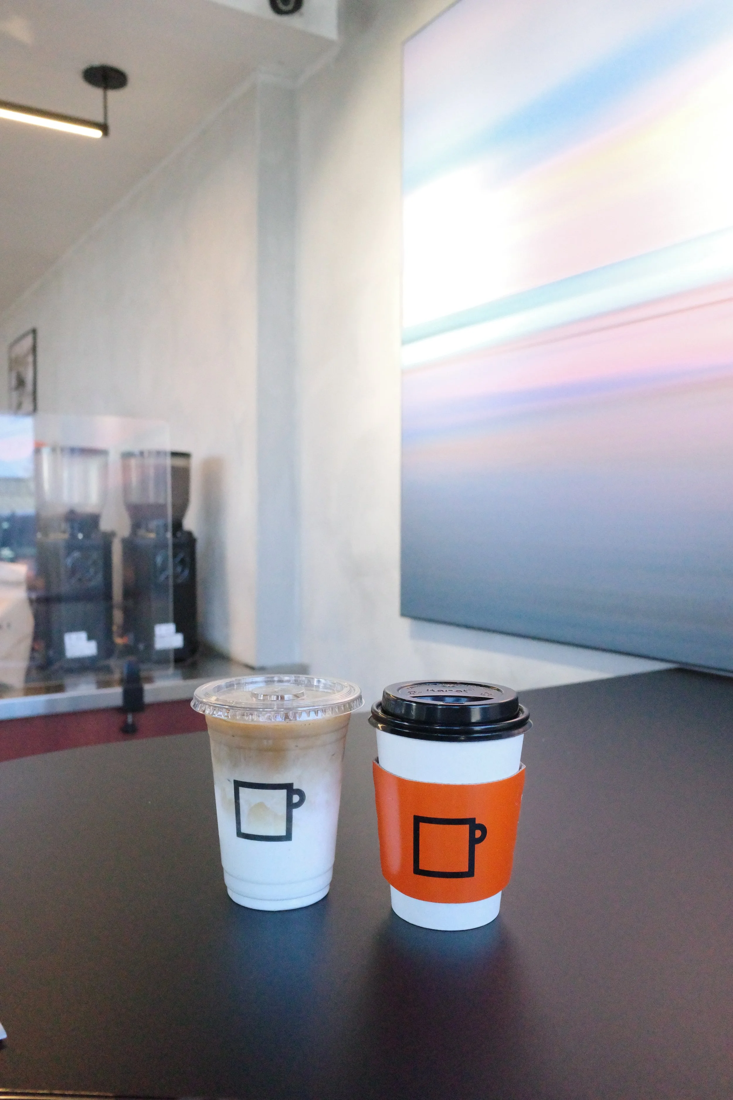 Two coffee cups on a table, one with a white sleeve and the other with an orange sleeve, both with black lids. In the background, there are coffee vending machines, a large colorful abstract wall art, and part of a ceiling with light fixtures. The se