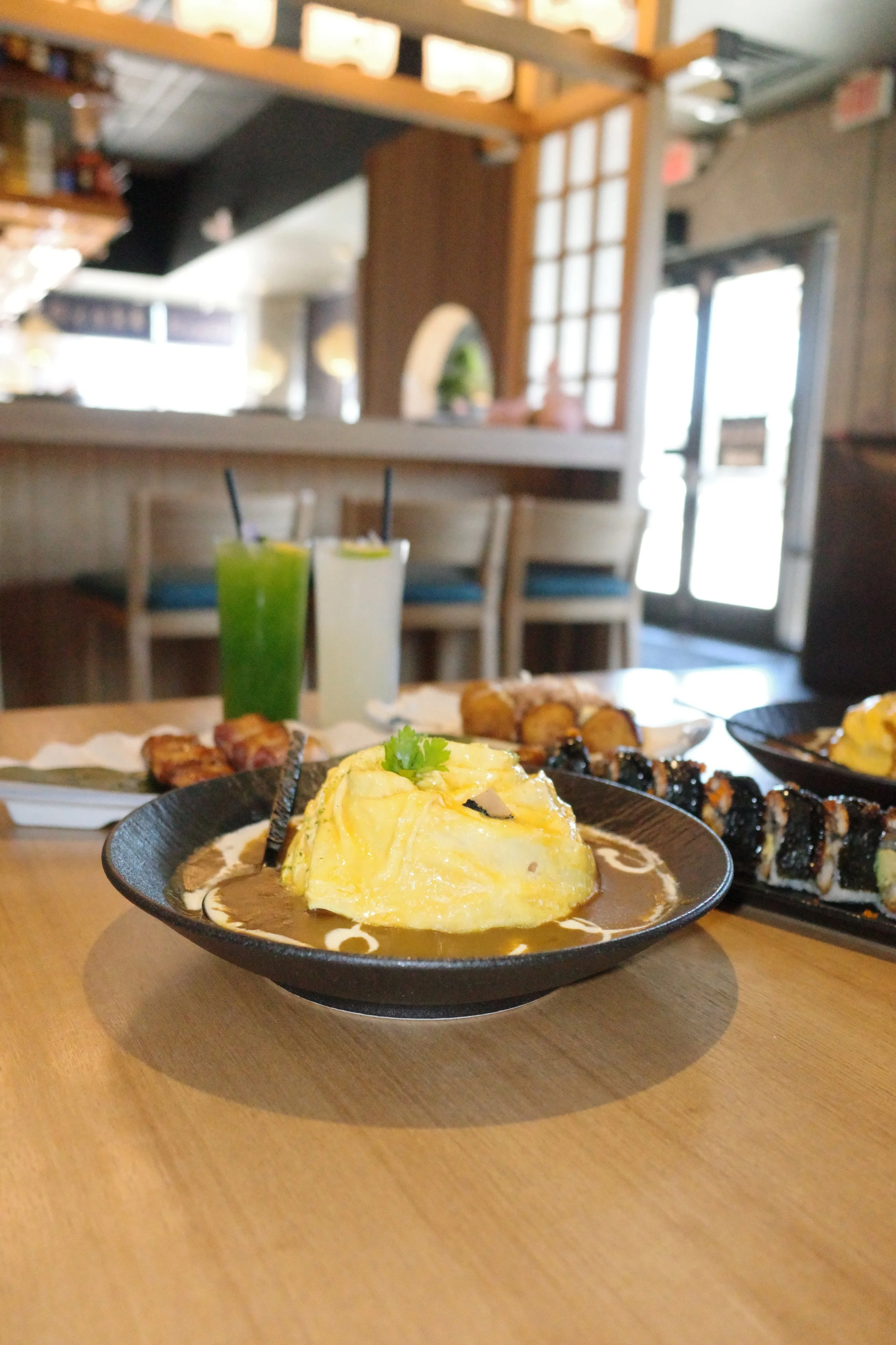 A table with various sushi dishes, including a plate with an omelet topped with cilantro, and drinks in tall glasses, in a restaurant setting with wooden chairs and bright natural lighting.