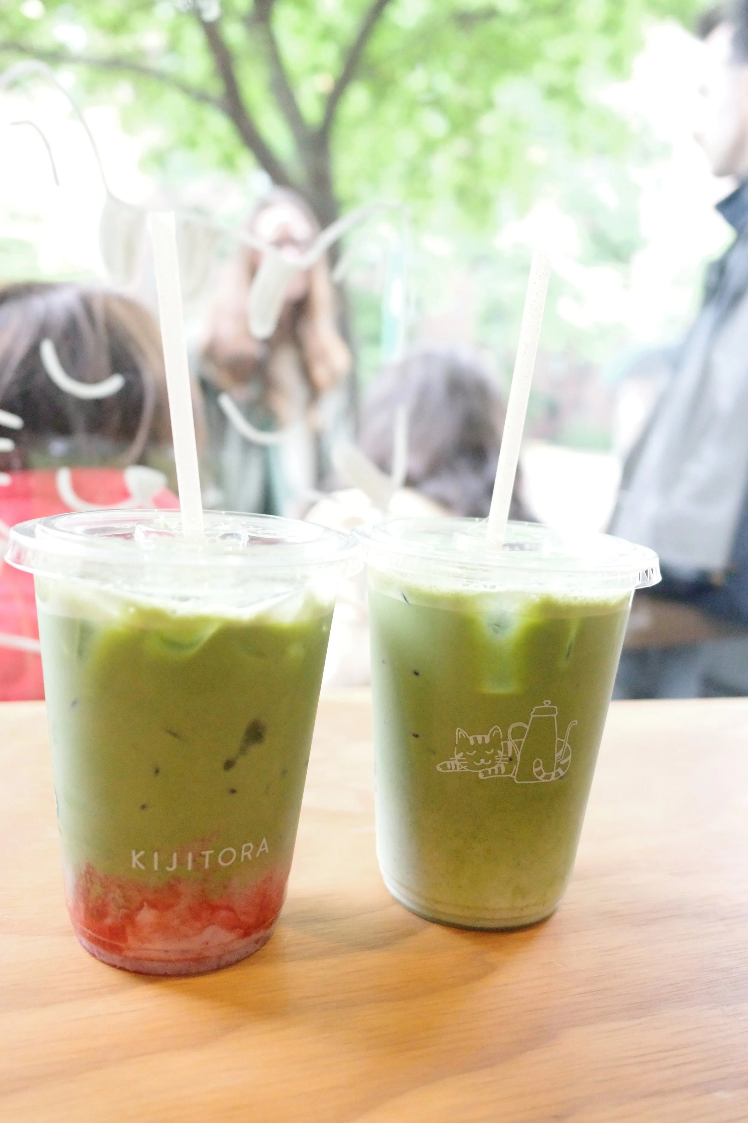 Two plastic cups filled with green smoothie with a white straw each, placed on a wooden table, with a blurred background of people and trees outside.