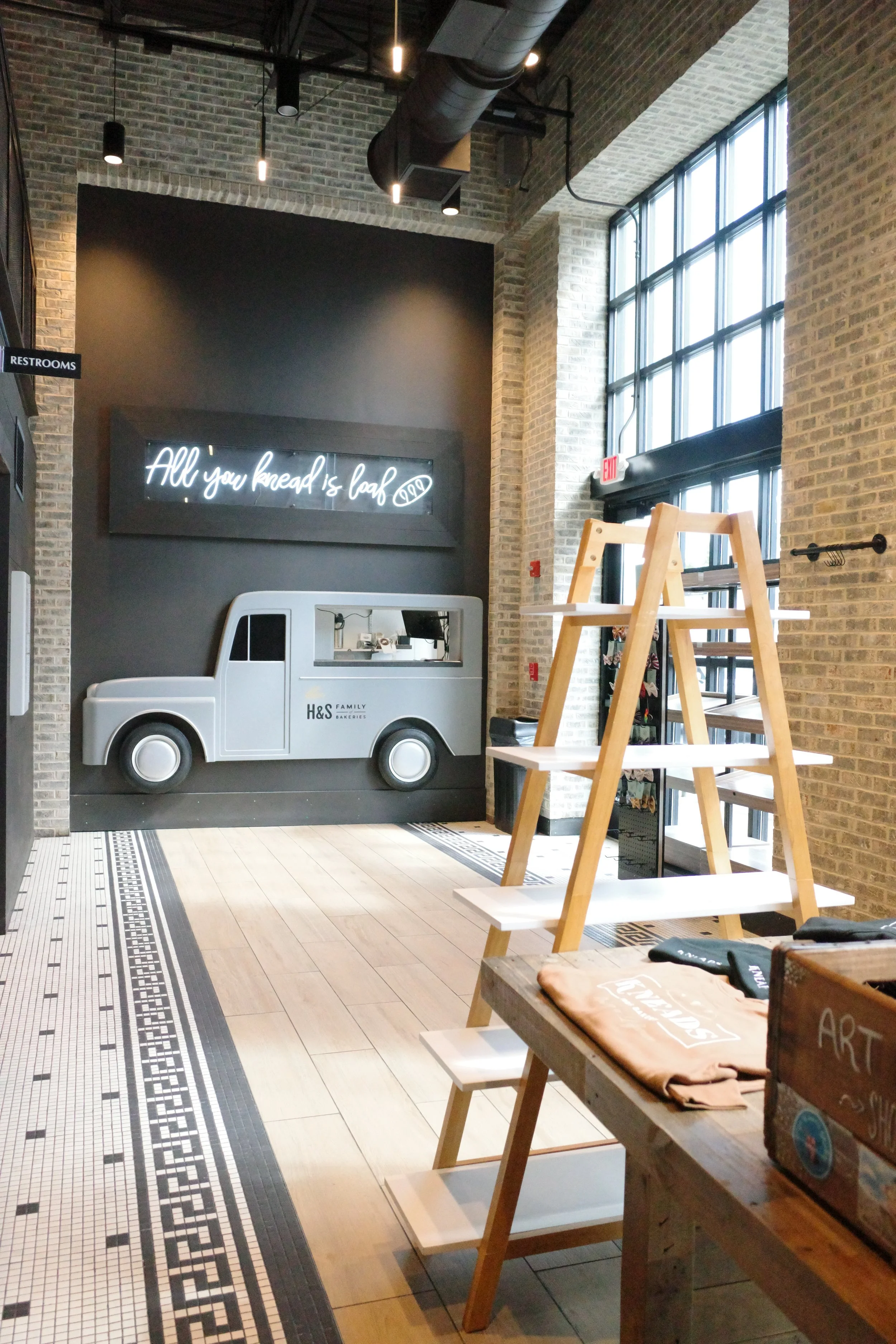 Interior of a bakery or café with a decorative mural of a truck on the wall, a neon sign that reads "All you bread is loaf," wooden shelving, a large window letting in natural light, and a tiled floor.