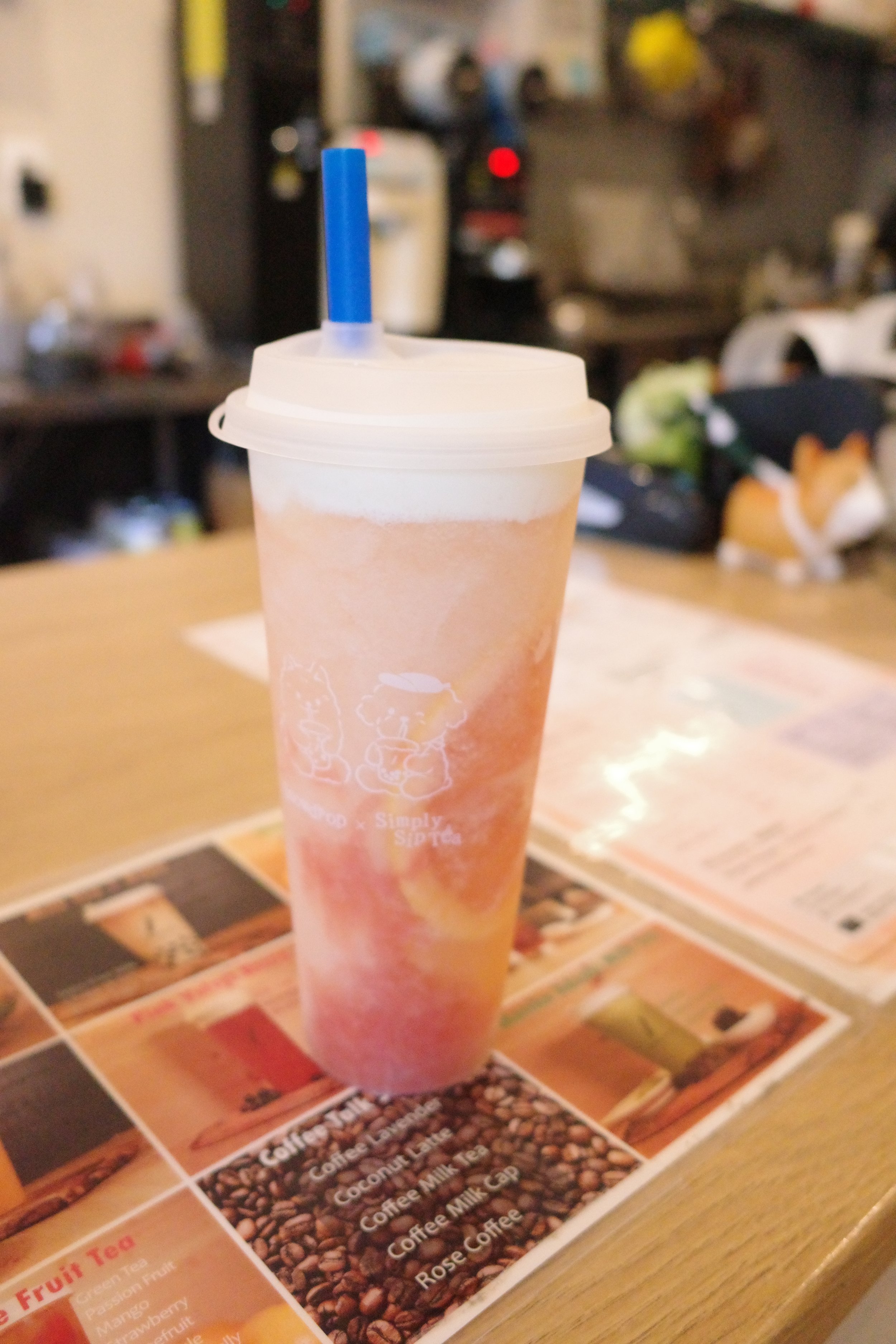 A paper cup with a plastic lid and a blue straw, placed on a menu or placemat with beverage options, with a blurred background of a restaurant or cafe interior.