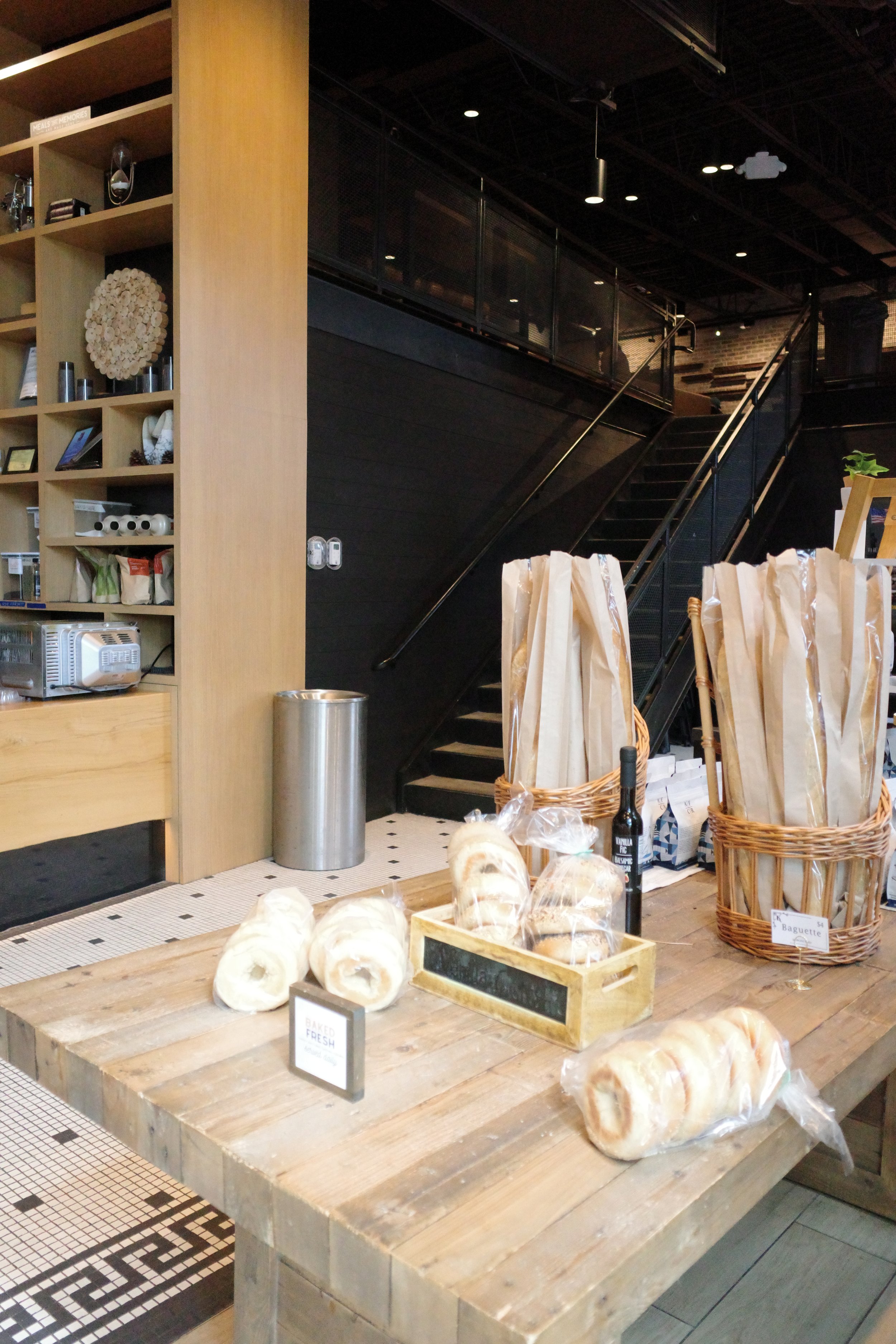Display of baked goods, including bagels wrapped in plastic on a wooden table, with baskets and a small chalkboard sign labeled "Baguette".