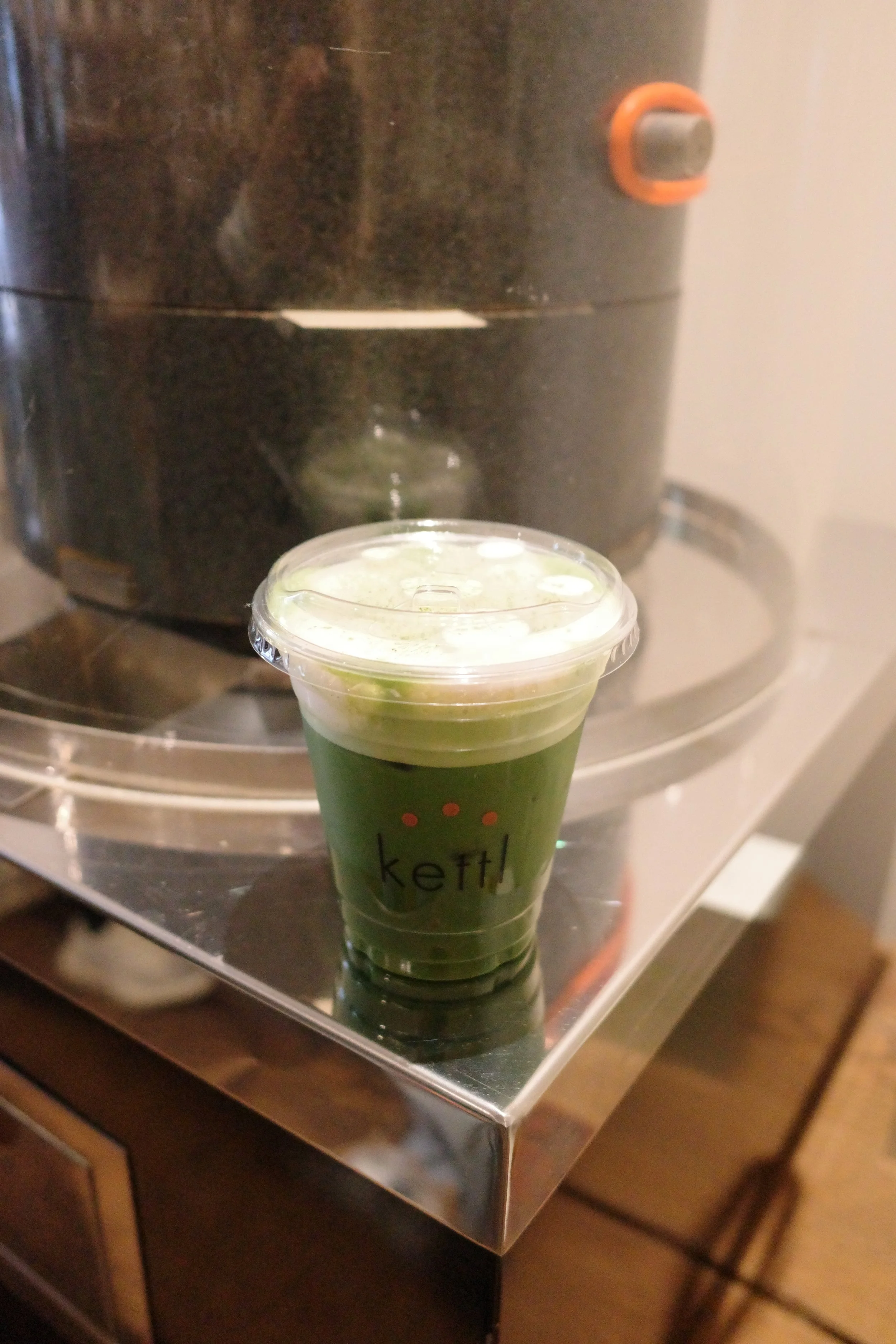 A green drink in a clear plastic cup with a transparent lid, sitting on a stove or hot plate. The cup has the word 'kett' partially visible and a small fruit or garnish on top.