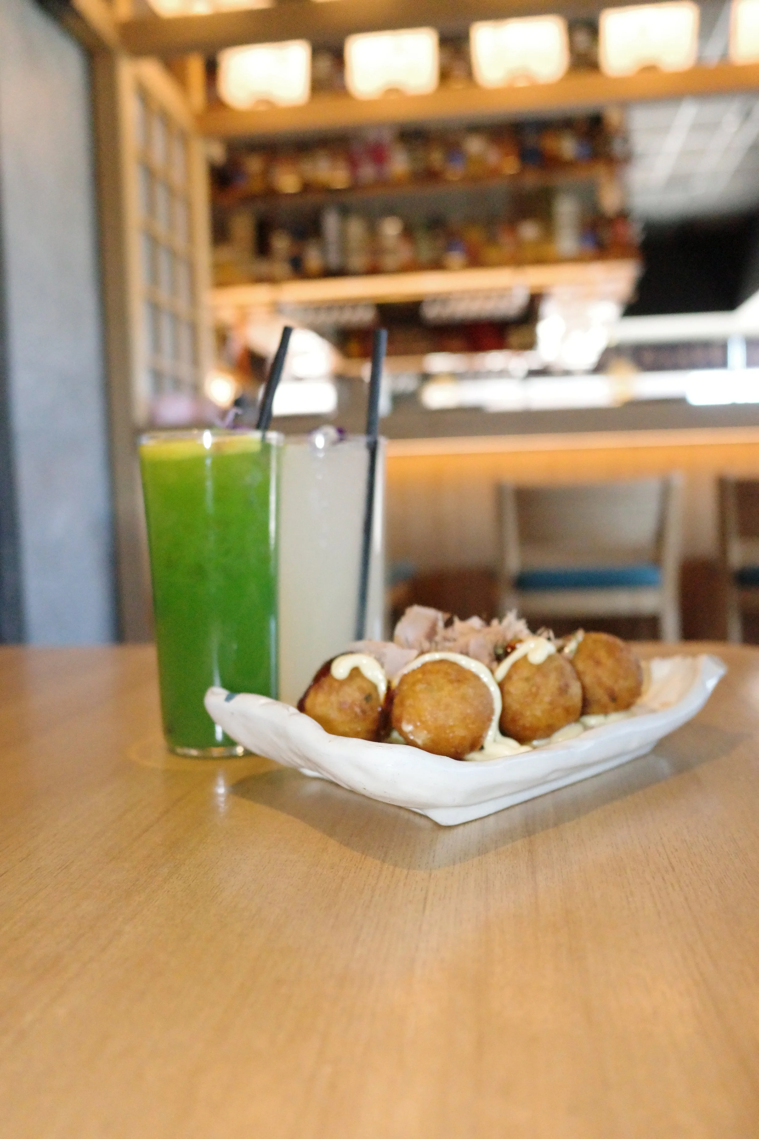 A plate of fried food topped with mayonnaise, with two colorful drinks, one green and one white, with black straws, on a wooden table in a restaurant or cafe setting.