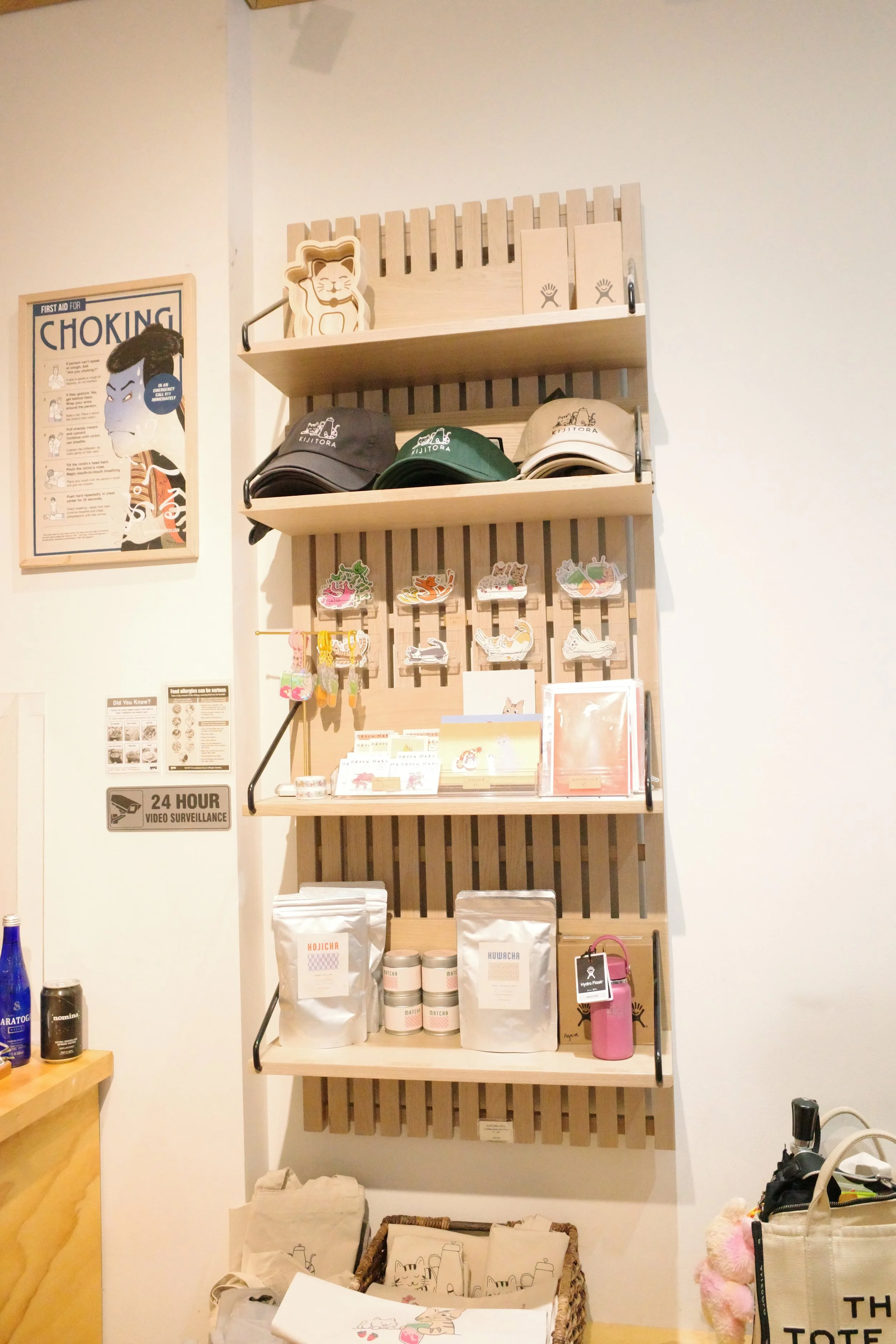 A wall-mounted wooden shelf with three levels displaying hats, stickers, and bags. There is a poster about choking on the wall to the left, and various items like bags and containers are visible below.
