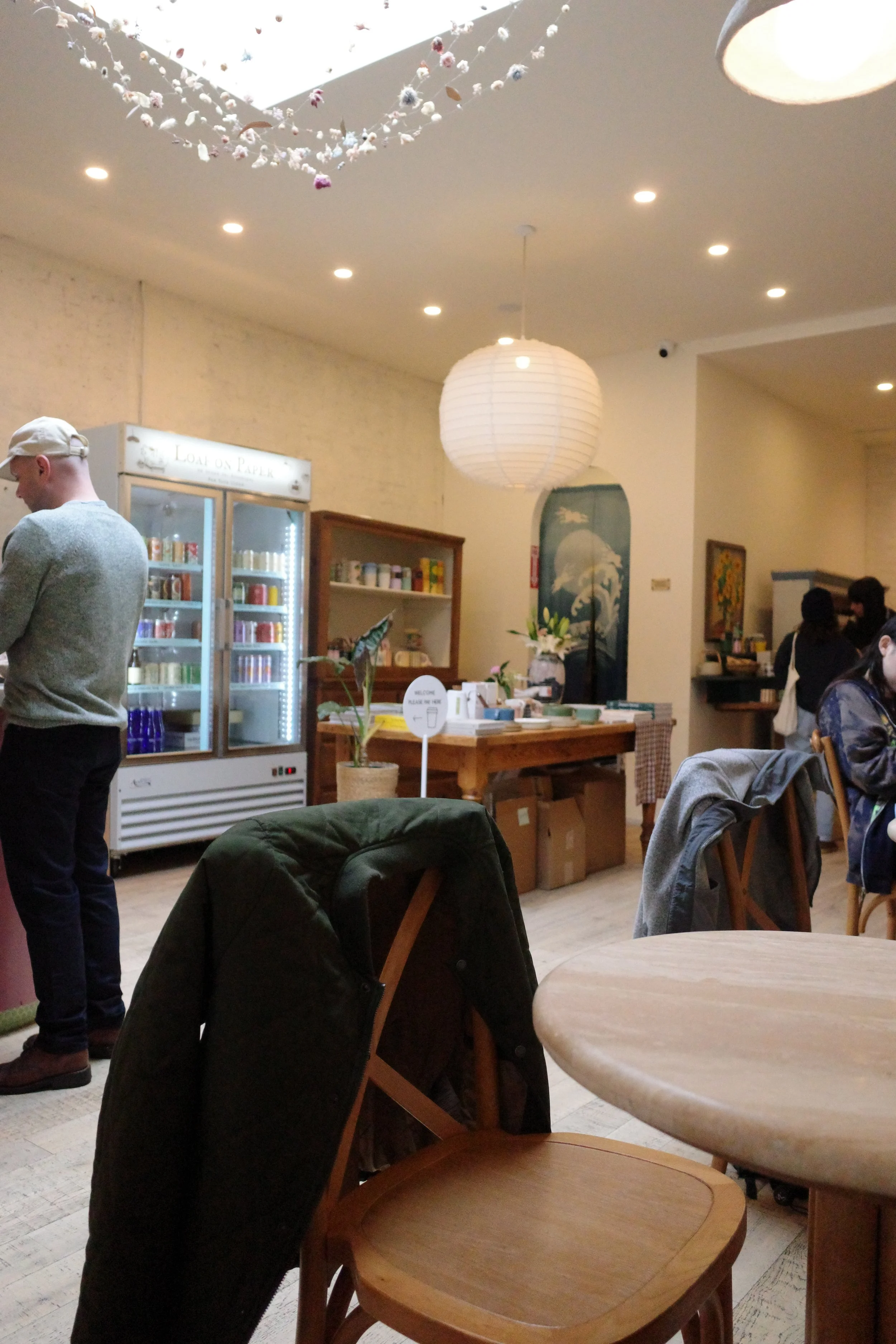 Inside a cozy cafe with wooden furniture, a person wearing a white cap and gray sweater stands near a glass refrigerator filled with beverages. The cafe features a paper lantern hanging from the ceiling, a mural or painting of a wave on the wall, and